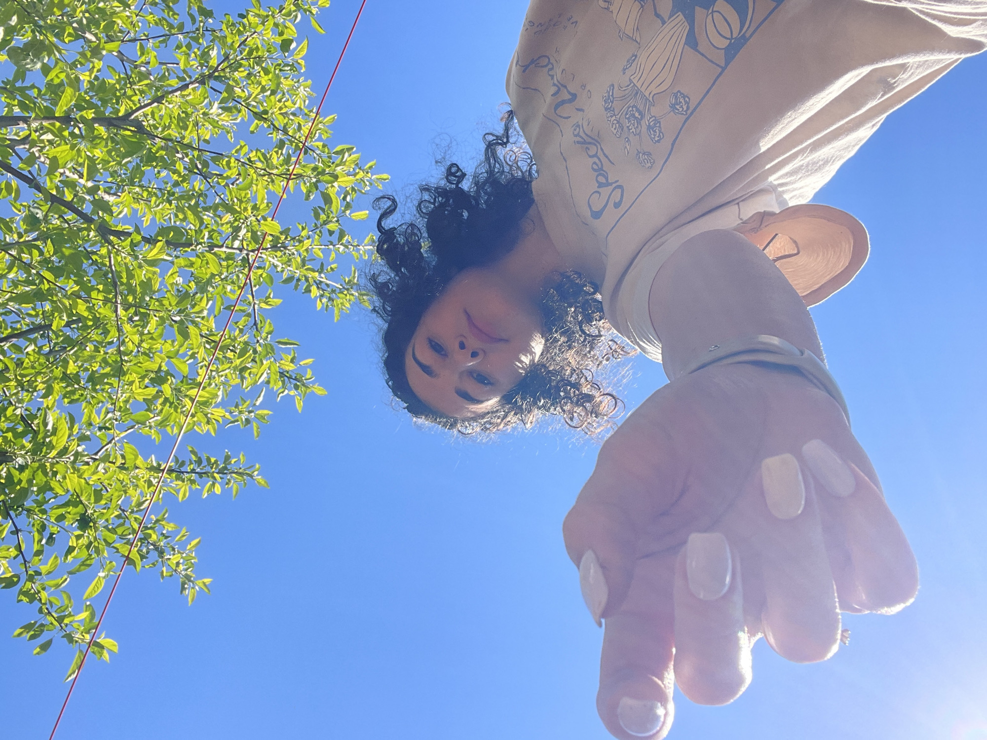 A low-angle, upward-facing photograph shows a person with curly black hair and tan skin. They are wearing a beige t-shirt with graphics. Their hand is extended towards the camera, showcasing manicured nails. A green tree branch is in the upper left corner. The background is a clear blue sky. The photograph was taken outdoors, likely in Sofia, Bulgaria.