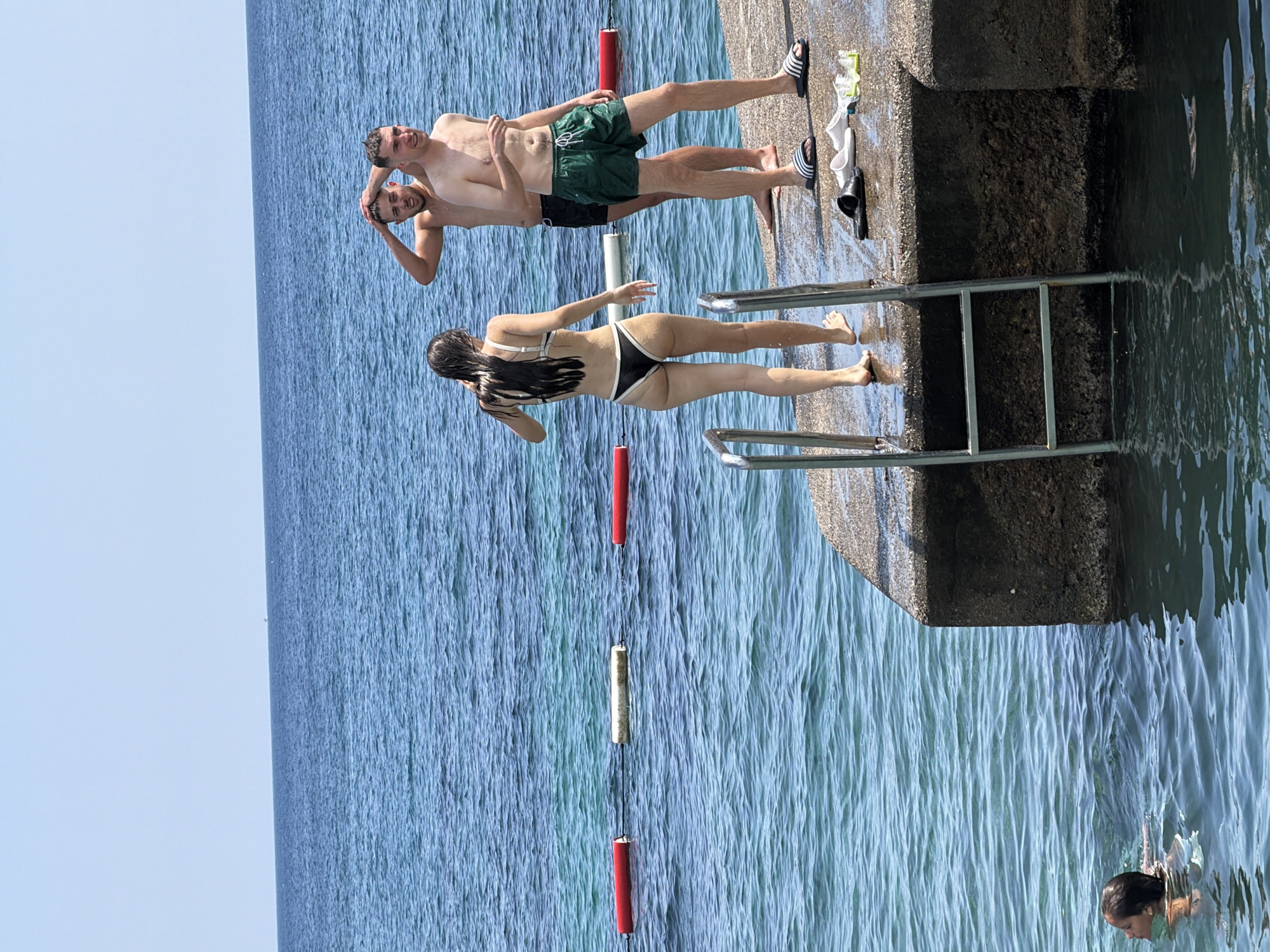 A group of four people are at a concrete pier on a clear, sunny day. The ocean is a vibrant blue with a few buoys visible in the water.
In the foreground, on the right side of the pier, a young woman in a black and white bikini stands facing away from the camera. Her hair is wet and dark. A metal ladder is attached to the pier to access the water. A young man in green swim trunks stands to her right, facing the water, with his left arm raised to his head. Behind him and slightly to his left, another young man, shirtless, stands with his right arm raised to his head, looking toward the first young man. Several pairs of flip-flops and goggles are scattered on the wet pier.
In the background, on the left side of the frame, a child with dark hair is swimming in the water. The horizon shows a faint line of land and a clear blue sky. The scene appears to be at a swimming area in Budva, Montenegro, likely near a beach or a popular spot for entering the sea.