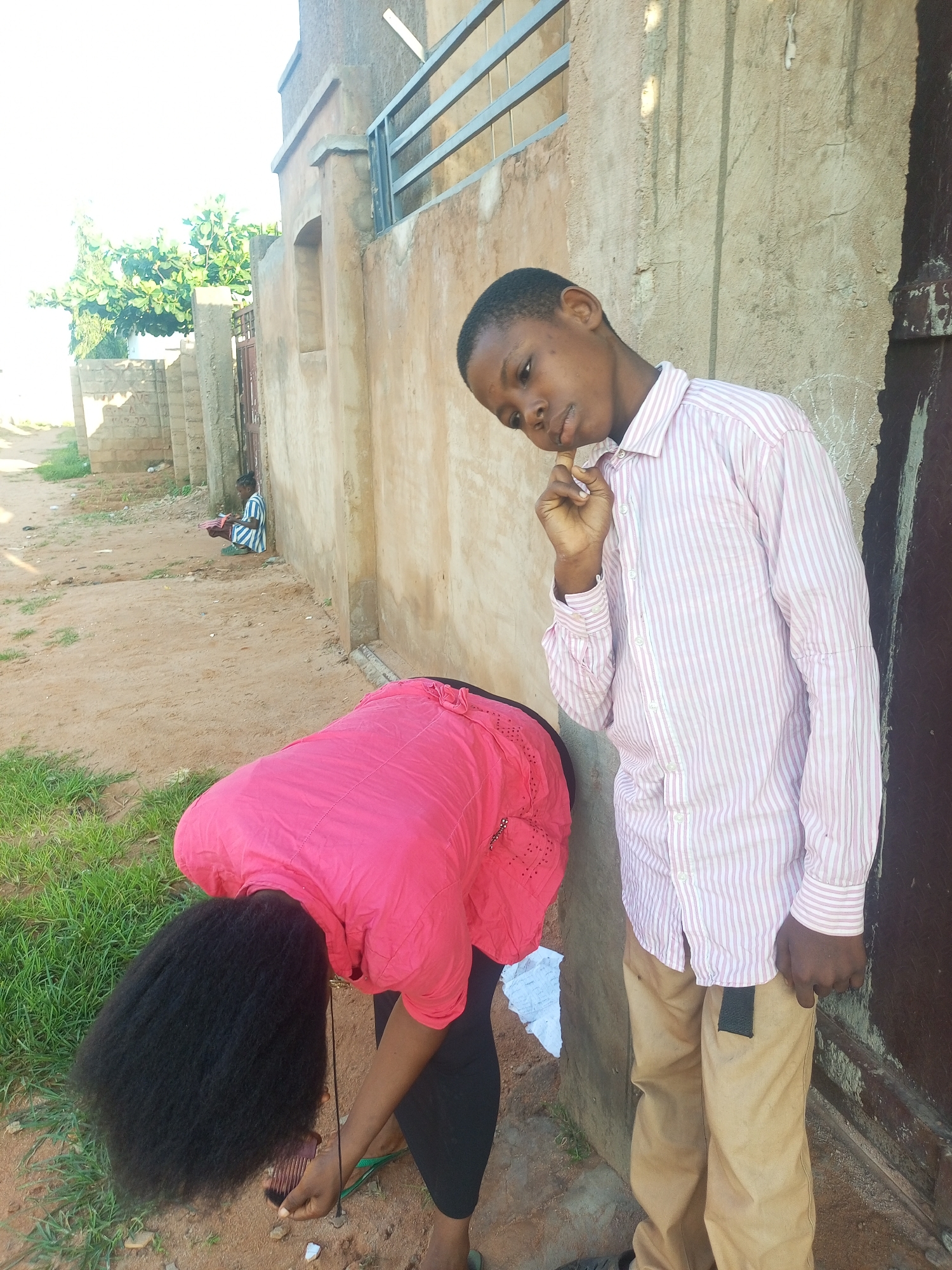 In Tempure, Nigeria, a young boy in a pink and white striped shirt and tan pants stands next to a textured, light brown wall. He is looking down and to the left, with his right hand near his chin. In the foreground, a person wearing a bright pink top and black leggings is bent over, tending to their long, dark, curly hair. A black comb is visible near their hand. In the background, further down a dusty path, another person is sitting on the ground, facing away from the camera and wearing a blue and white striped garment. The environment appears to be an outdoor setting with some grassy areas and a rough wall bordering a pathway.