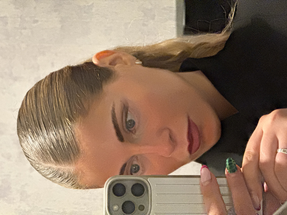 The image displays a close-up mirror selfie of a woman within an indoor environment. The woman, appearing to be in her twenties or thirties, is positioned slightly off-center to the left, directing her gaze towards the smartphone held in her left hand, which is visible in the lower-left quadrant of the frame. Her light-colored hair is slicked back tightly with a distinct center part, and some wavy, blonde strands extend over her right shoulder. She exhibits a warm skin tone, light blue eyes, dark, well-defined eyebrows, and black winged eyeliner. Her lips are painted with a dark red or plum-colored lipstick. She is attired in a dark garment, likely black, with a high collar. A small, silver-toned hoop earring is visible on her left ear. On her left ring finger, a silver-toned ring with a rectangular facet is present. Her left thumbnail features a pink French manicure, while her left index finger displays a textured, dark green nail design. The smartphone, consistent with an iPhone model due to its three-lens camera array, is encased in a ribbed, light beige protective cover. The background is a light grey wall with a subtle, mottled texture. Based solely on the visual information provided, there are no identifiable cues within the image to determine a specific location within the city of Paris, France.