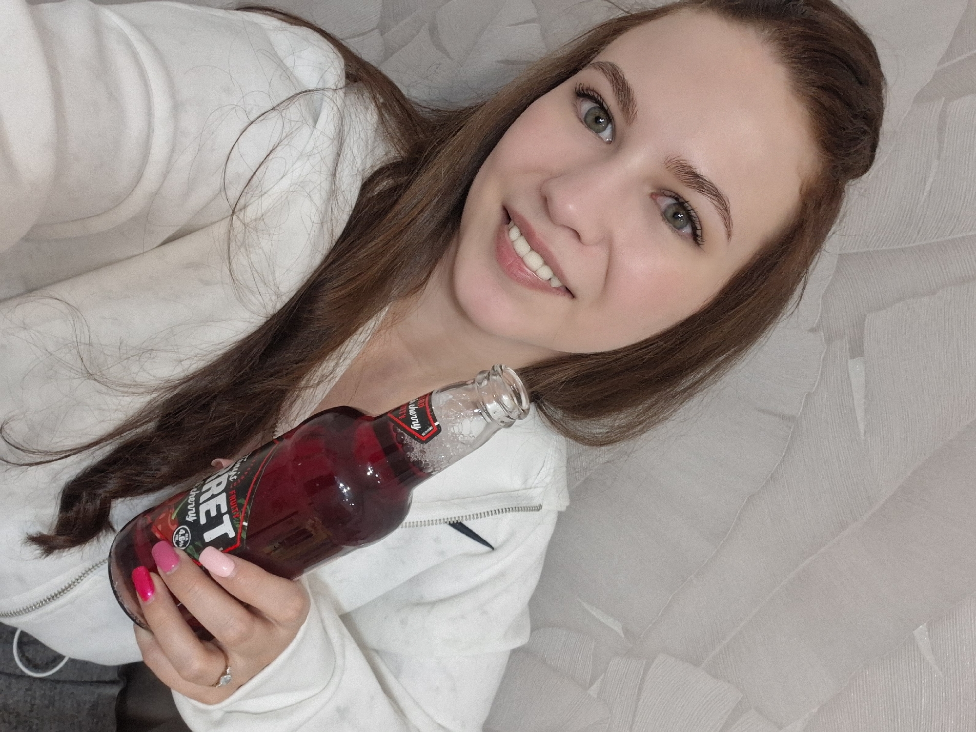 A fair-skinned woman with long brown hair and green eyes, wearing a white zip-up hoodie, is lying on a light grey textured surface, possibly a sofa or bed. She is holding a bottle of cherry-flavored alcoholic beverage called "Garretts." Her nails are painted in alternating shades of pink. She is smiling at the camera. The exact location in the city of Chaikovsky, Russia cannot be determined from the image.