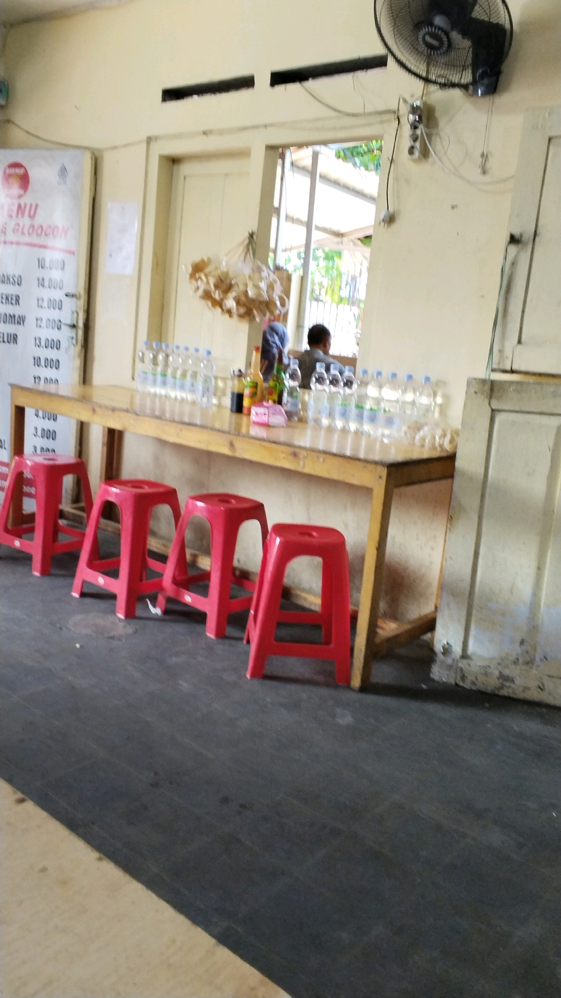 The image depicts an indoor scene, likely a restaurant or eatery, with a long wooden table as the central focus. Four bright red plastic stools are arranged in a row in front of the table. On the table, numerous bottles of water are neatly lined up, along with a few condiment bottles and some food items hanging from the ceiling. In the background, through an open doorway or window, greenery is visible, suggesting an outdoor area beyond. Two individuals are partially visible behind the table, facing away from the camera. One person appears to be wearing a head covering. The walls are painted a pale yellow, and a ceiling fan is mounted on the wall. A menu board with prices in Indonesian Rupiah is displayed on the left side of the image. The floor is a dark, textured surface. The overall atmosphere suggests a casual dining establishment. The text on the menu board, visible in the image, mentions "MENU" and "GLOGON" and lists prices for various items like "BAKSO," "SIOMAY," and "TELUR." The presence of Indonesian currency and text indicates the location is likely in Indonesia. The specific location of "Sooko" is not explicitly mentioned in the visual data but contextually could be a district or area within Indonesia where such an establishment might be found.