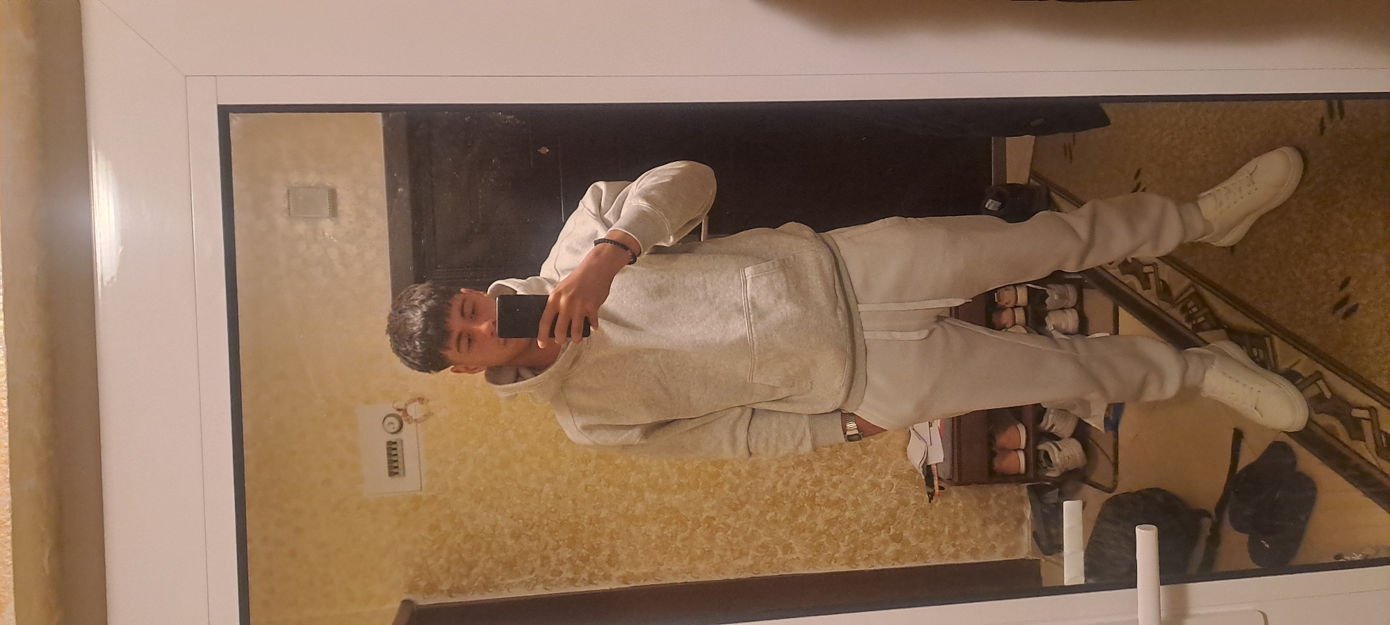 A young man takes a selfie in a mirror. He is wearing a light gray hoodie and sweatpants, along with white sneakers. Behind him, there is a dark door and a patterned wall. Below him are shoes stored in a rack, along with some personal items. It's not possible to determine the location in Burgas, Bulgaria from this image.