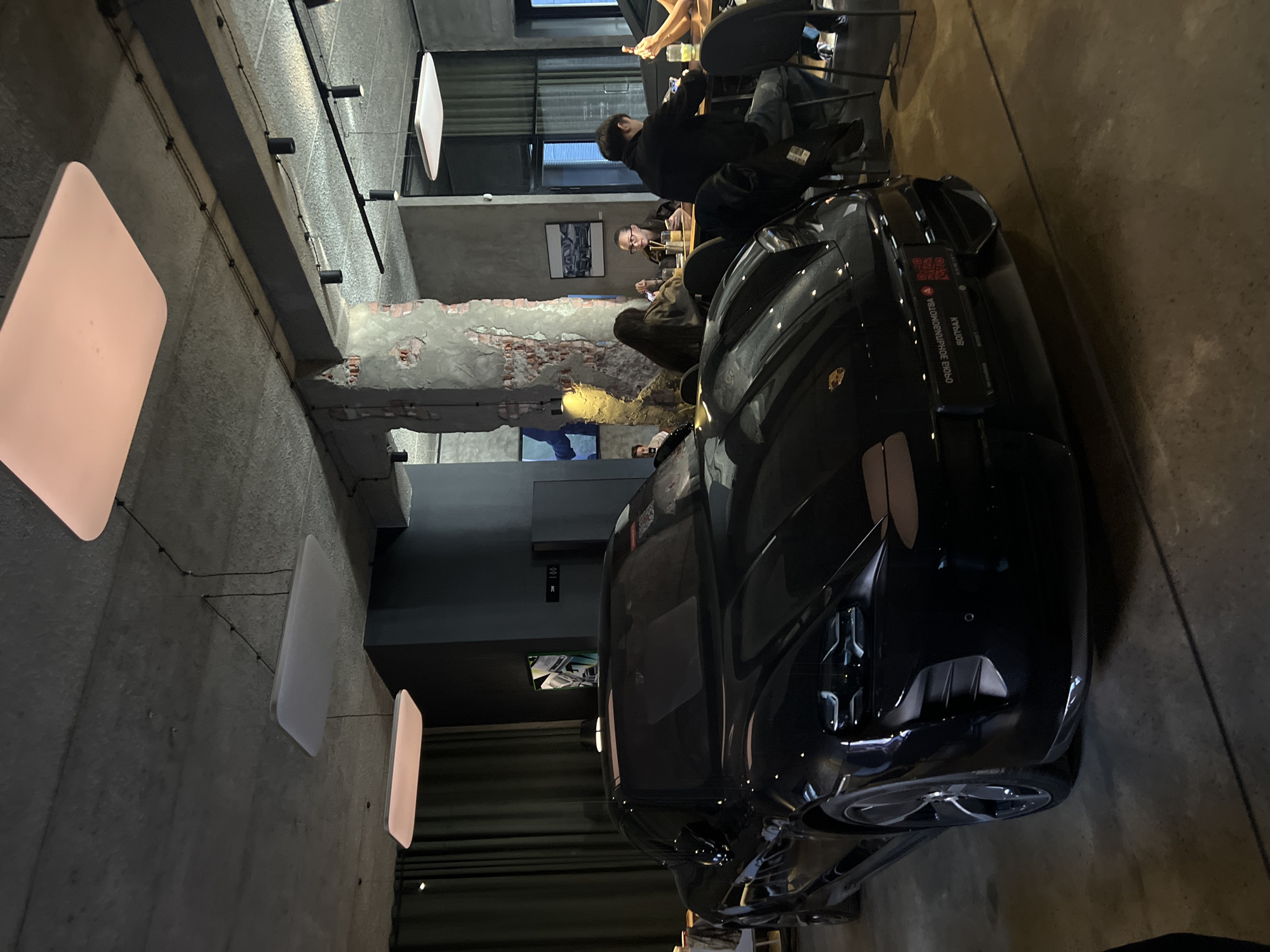 The photograph depicts an interior scene, seemingly a restaurant or cafe, with modern architectural elements and a casual, relaxed atmosphere.
**Location:** The car has a QR code on the windshield that says "BOTRAN" and appears to be part of a promotion. BOTRAN is a rum distillery based in Guatemala. The QR code has an associated website, which could lead to Saint Petersburg, Russia.
**People:** Several individuals are present, scattered around tables in a dining area. Some are seated, appearing to be engaged in conversation or consuming food/drinks. Others are milling around or moving. The lighting is subdued, creating shadows that obscure some of their features.
**Objects:** Dominating the foreground is a dark-colored sports car, likely a Porsche, angled towards the viewer. Its sleek design and reflective surface are prominent. Beyond the car, the interior consists of tables, chairs, and what appears to be a counter area in the background. There are rectangular ceiling-mounted light fixtures, adding to the ambiance. The walls have a textured appearance, possibly brick or stone, adding to the overall modern aesthetic.
**Actions:** The people in the scene are engaged in typical restaurant activities - eating, talking, and socializing. The car's presence seems to be more of a display piece or an advertisement of some sort.
**Interactions:** The focus is on the general ambiance and the car, rather than specific interactions between individuals. There is a sense of casual interaction and social activity, typical of a restaurant setting.
**Environment:** The interior is modern, with a mixture of industrial and stylish design elements. The walls are a mix of exposed brick and concrete. The lighting is a combination of natural light from the windows and artificial light from the light fixtures. Overall, the environment is designed to be casual and inviting.
