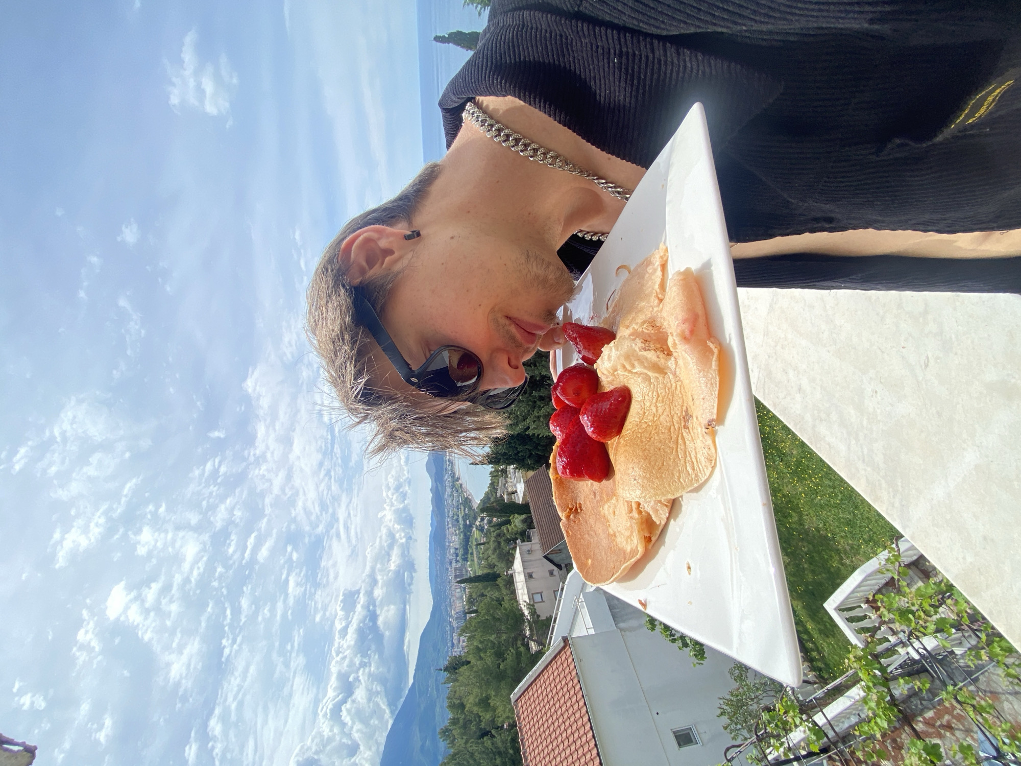 The image, originally oriented on its side, captures a bright outdoor scene featuring a person about to eat, set against a panoramic coastal backdrop.
In the foreground, a young individual, appearing male, with light brown hair and subtle facial stubble, is seen in profile leaning downwards towards a white rectangular plate. He wears dark sunglasses, a black stud earring, and a prominent silver cuban link chain necklace. His attire consists of a dark, textured top, possibly corduroy. His attention is focused on the food directly in front of him.
On the white plate are several golden-brown pancakes or crepes, generously adorned with fresh, vibrant red strawberries. The setting suggests an outdoor dining experience, likely on a patio or balcony, with part of a light-colored table surface visible.
The background unfolds into a scenic vista under a clear blue sky with scattered white clouds, indicating a sunny day. Below the sky, a mountainous landscape is visible, featuring lush green vegetation, numerous trees, and residential buildings with characteristic red-tiled roofs. In the distance, a body of water, identified as the Adriatic Sea, stretches along the horizon, bordering the coastal town of Šušanj, Montenegro. The overall atmosphere is tranquil and inviting, depicting an enjoyable outdoor meal with a beautiful view. No visible text is present in the image.
