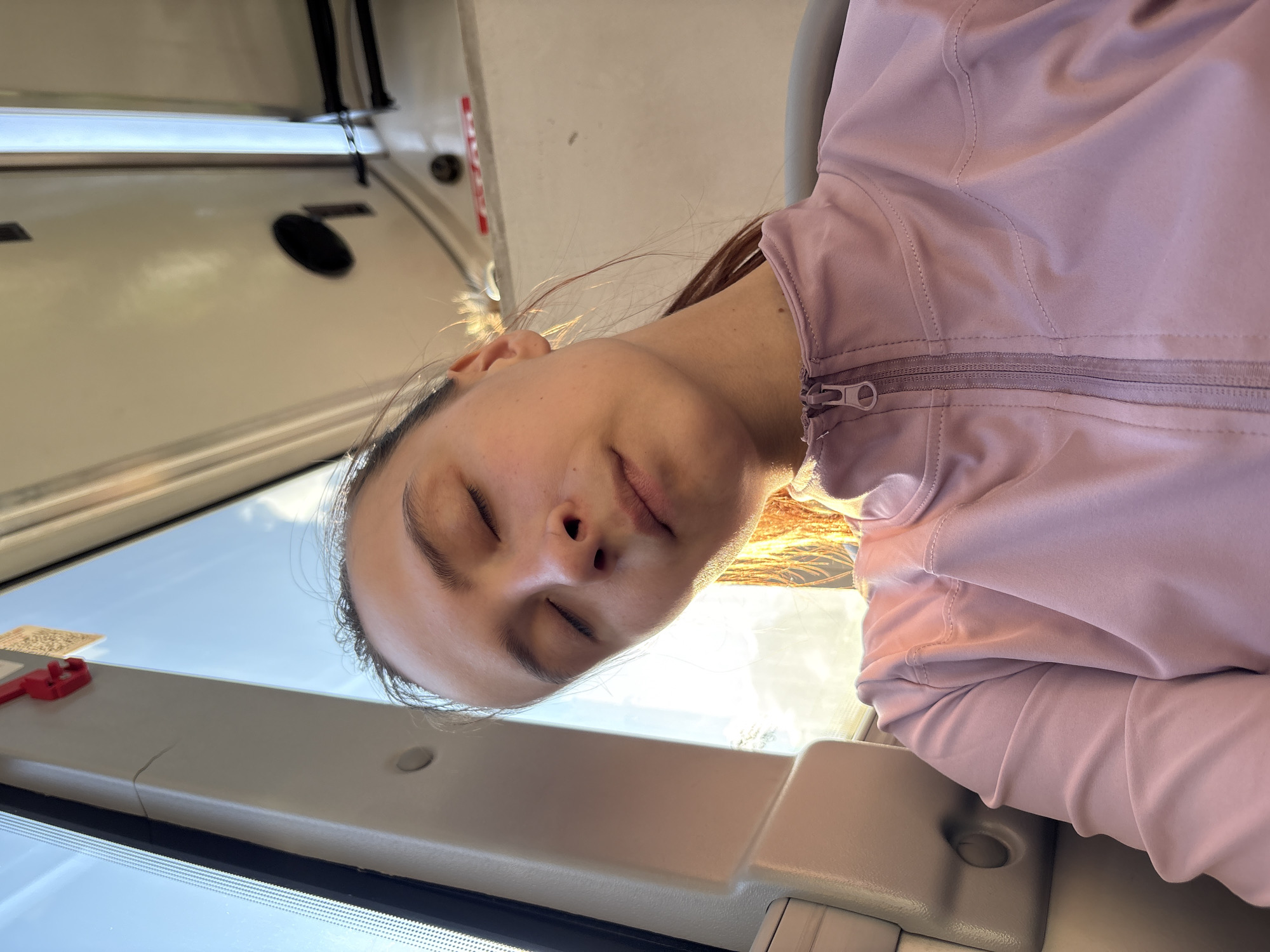 A young woman is resting, possibly sleeping, on what appears to be public transportation. She is wearing a light pink jacket. The background suggests she is near a window with a view of a blue sky, indicating daytime. The interior elements of the vehicle are visible, but specific details like the type of transportation are not clear. There is no information in the image to suggest the location is Perm, Russia.