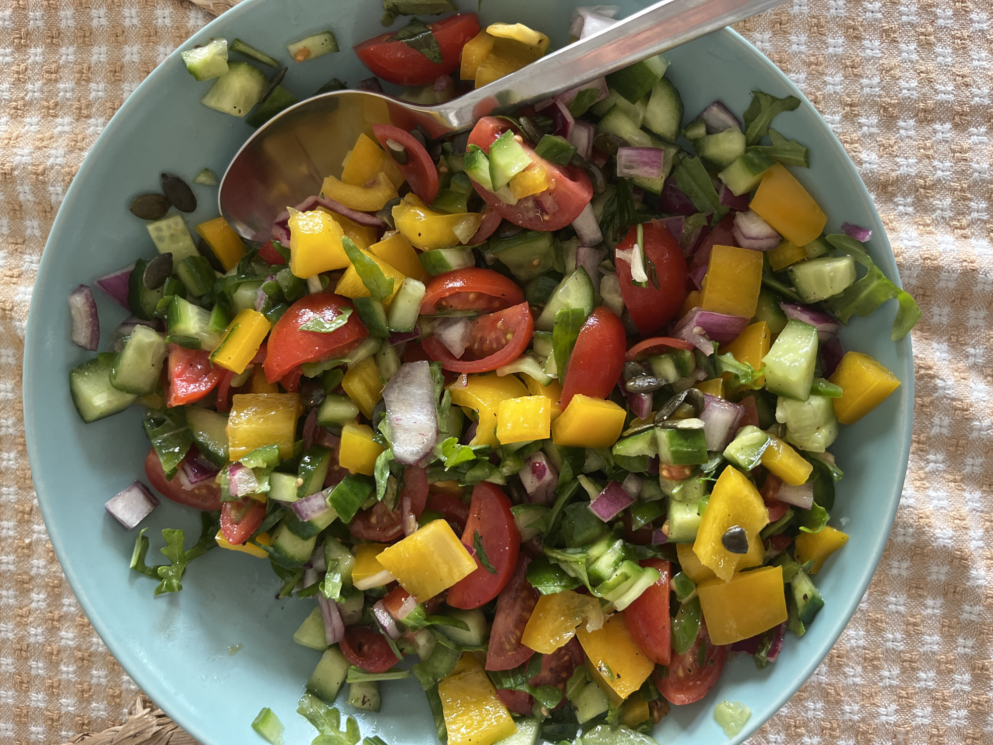 A mixed salad is contained within a light blue, circular bowl, presented from a top-down perspective. The salad comprises chopped red tomatoes, diced green cucumbers, cubed yellow bell peppers, sliced red onions, and various green leafy components. Pumpkin seeds and small dark objects, likely capers, are distributed throughout the mixture. A silver-colored metal spoon is positioned partially within the salad on the bowl's right side. The bowl rests upon a light orange and tan woven tablecloth displaying a subtle grid pattern. This scene is located in Ramat Gan, Israel.
