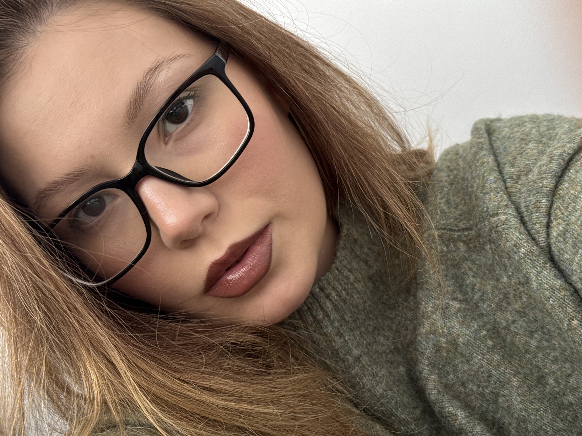 A close-up selfie of a fair-skinned woman with light brown hair. She is wearing black-framed glasses and a muted green, possibly knitted, sweater. Her lips are colored with a lipstick in shades of brown and pink. The background appears to be a plain white wall. The location is Unknown, Unknown.