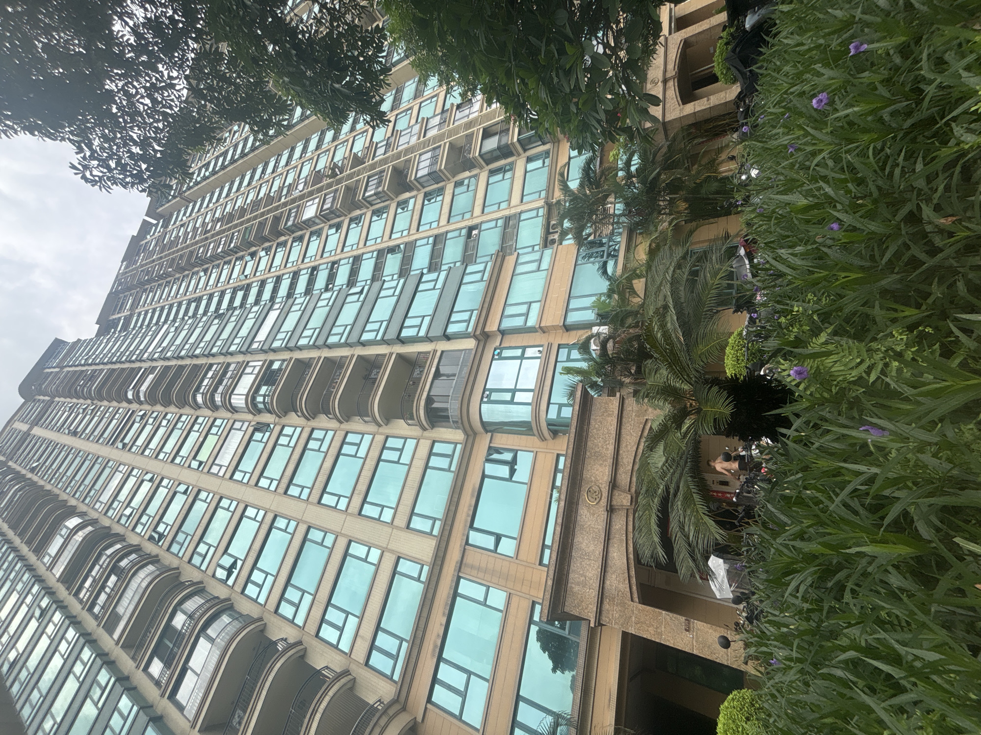 The image shows a very tall, modern residential building in Nanhai District, China, with a facade of beige stone and numerous light blue tinted windows. The building has a repeating pattern of balconies with black railings on alternating floors. In the foreground, there is lush green vegetation with scattered purple flowers. Several motorcycles are parked in front of the building, behind the foliage. A person, appearing to be a male with no shirt, is visible standing near the motorcycles and the entrance of the building. The sky above is overcast.