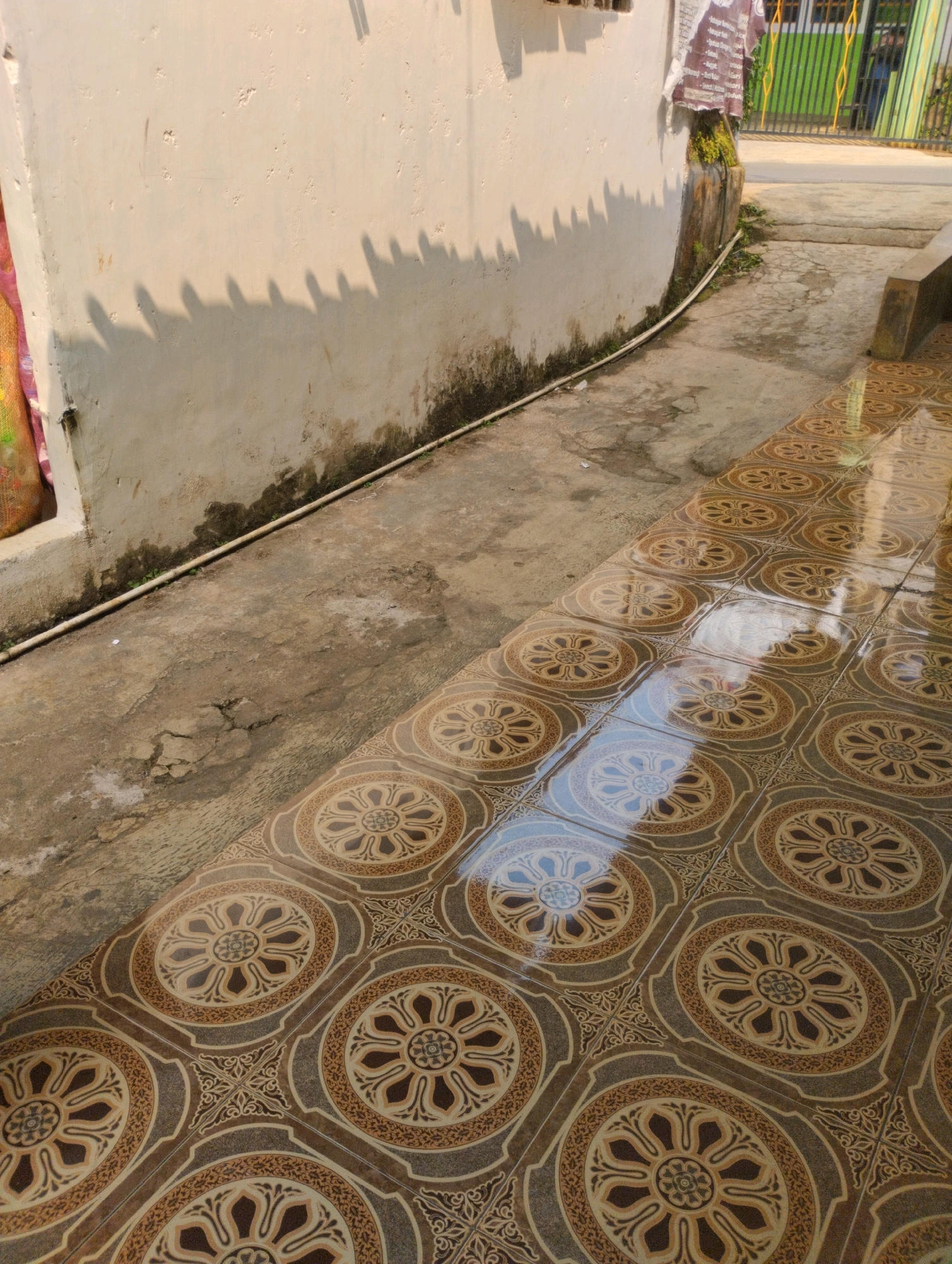 This is a high-angle, medium shot of a pathway with patterned tiles in Cimenyan, Indonesia. The foreground is dominated by a section of floor covered in brown and tan square tiles, each featuring a circular, floral-like design with intricate patterns. The tiles are wet and reflective, showing a bright sky and the white wall casting shadows.
To the left of the tiled path, there is a concrete walkway. A section of a textured, off-white wall runs along the edge of the concrete. The lower part of this wall has some dark staining and moss. A white cable runs along the base of the wall and then across the concrete walkway. The concrete itself is cracked and weathered in places.
The background shows a green metal fence with vertical bars, and beyond that, some buildings and trees are visible. Sunlight creates distinct, somewhat jagged shadows from an unseen object on the white wall. A small portion of a colorful fabric, possibly part of a bag or clothing, is visible on the far left edge of the frame, near the wall. The overall scene appears to be an outdoor area in front of a building or house.