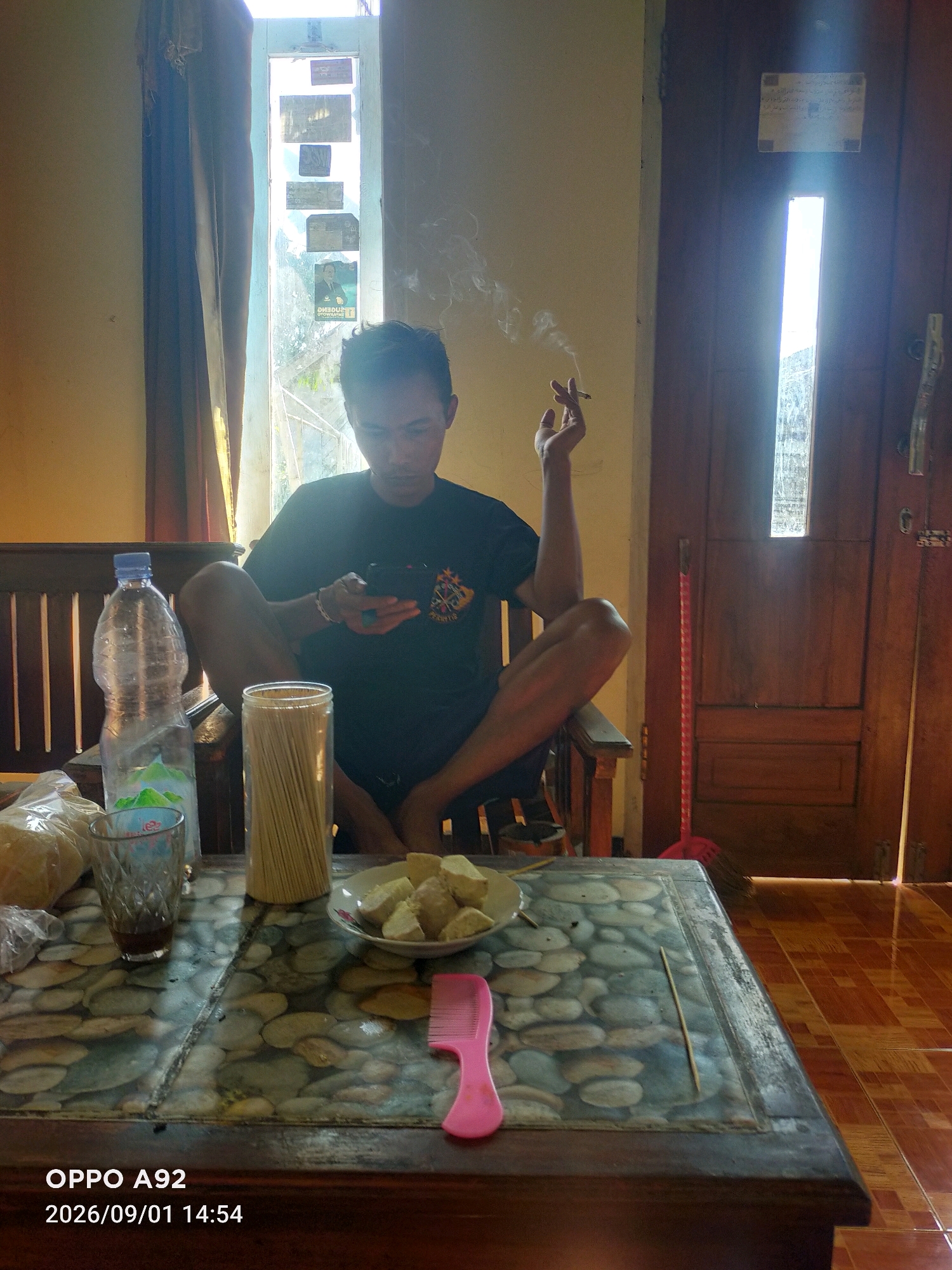 The image depicts a man sitting cross-legged on a wooden chair indoors, with a cigarette in his right hand, emitting smoke. He is wearing a black t-shirt and appears to be looking at his mobile phone, which he holds in his left hand. In front of him is a table with a patterned surface, on which are placed a bottle of water, a glass with a dark liquid, a container of wooden skewers, a plate of cubed food, and a pink comb. The background features a window with curtains and several papers or photographs affixed to it, as well as a wooden door with a vertical glass pane. A red broom leans against the door. The floor is tiled with brown square tiles. The lighting suggests it is daytime, with sunlight entering from the window. The timestamp "2026/09/01 14:54" and "OPPO A92" are overlaid at the bottom left of the image. The scene is likely in a residential setting in Binangun, Indonesia, given the timestamp's format and the typical presentation of such images.