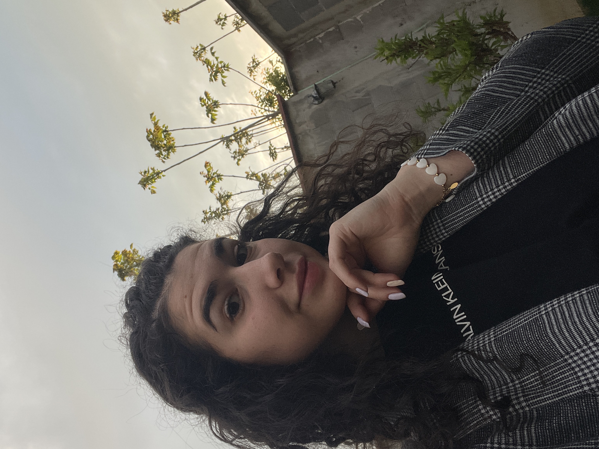 A young woman with curly dark hair is posing for a selfie outdoors. She is wearing a black Calvin Klein t-shirt and a grey and white plaid jacket. She is wearing a bracelet with white heart-shaped beads on her wrist. The sky is visible in the background, along with the branches of a tree with green leaves. A concrete building is also visible. The location may be Sofia, Bulgaria.