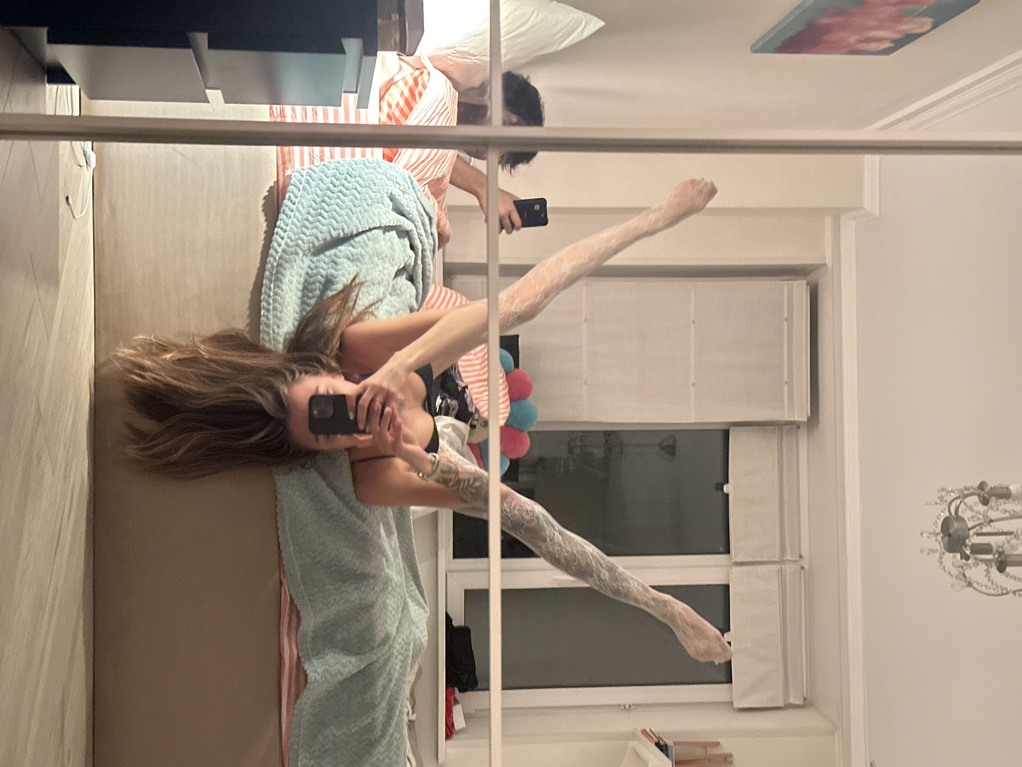This is a full-body, slightly angled, candid shot of a young woman lying upside down on a bed, holding her phone to take a selfie. The woman has long, brown hair that cascades down past her head. She is wearing a black tank top. Her legs are raised in the air and appear to be wrapped in white bandages or a similar material. In the background, another person, appearing to be a young man, is visible sitting on the bed, looking at his phone. He has dark hair and is wearing an orange and white striped shirt. The bed is covered with a light blue textured blanket and a striped comforter. The room appears to be a bedroom with a large window covered by white blinds in the background. A chandelier is visible on the ceiling. The image is reflected in a mirror that frames the left side of the scene, showing part of a dresser and a framed picture on the wall. The overall impression is that of a casual, indoor setting, possibly a home or hotel room. The location is not specified in the image.