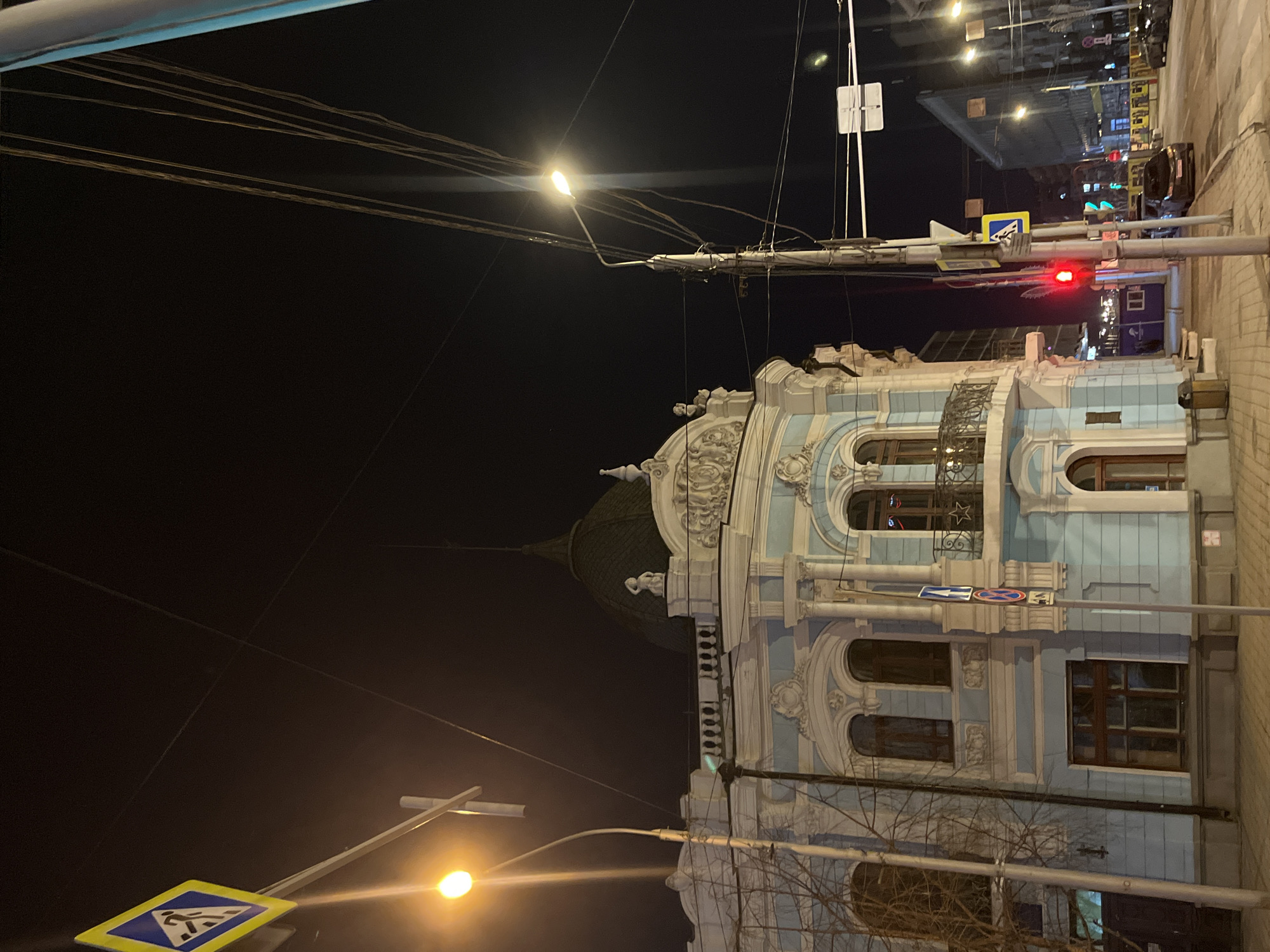 The image shows a night scene in Krasnoyarsk, Russia, featuring a historical building with distinct architectural details, including a dome and ornate facade. The building appears to be well-lit and maintained. Streetlights and electrical wires are visible in the foreground and above the building. Traffic lights are illuminated in red, and pedestrian crossing signs are present, indicating an urban environment with vehicular and pedestrian traffic. The street appears paved. The sky is dark, typical of a night scene. Trees are partially visible in the foreground.