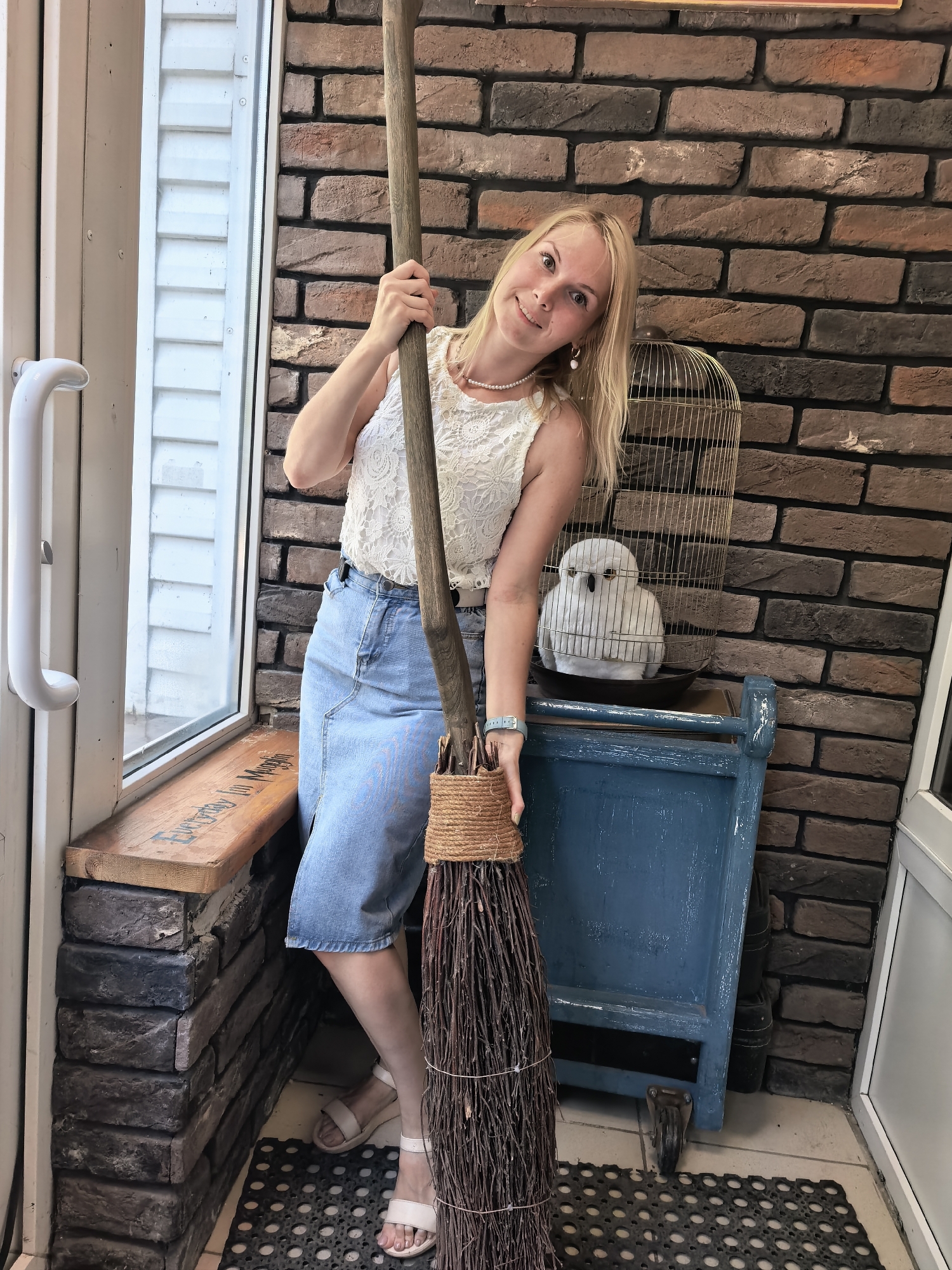 A blonde woman stands indoors, leaning against a broomstick that is propped up next to a blue cabinet. The woman is wearing a white lace top and a denim skirt. She has a pearl necklace and earrings. On the blue cabinet, there is a decorative birdcage with a white owl inside. The background is a brick wall. The scene appears to be a shop or a themed area, possibly in Sterlitamak, Russia, given the prompt. The floor is tiled and a black mat is placed in front of the cabinet. There is a window on the left with a white vertical siding and a wooden bench beneath it. The lighting is natural, coming from the window.