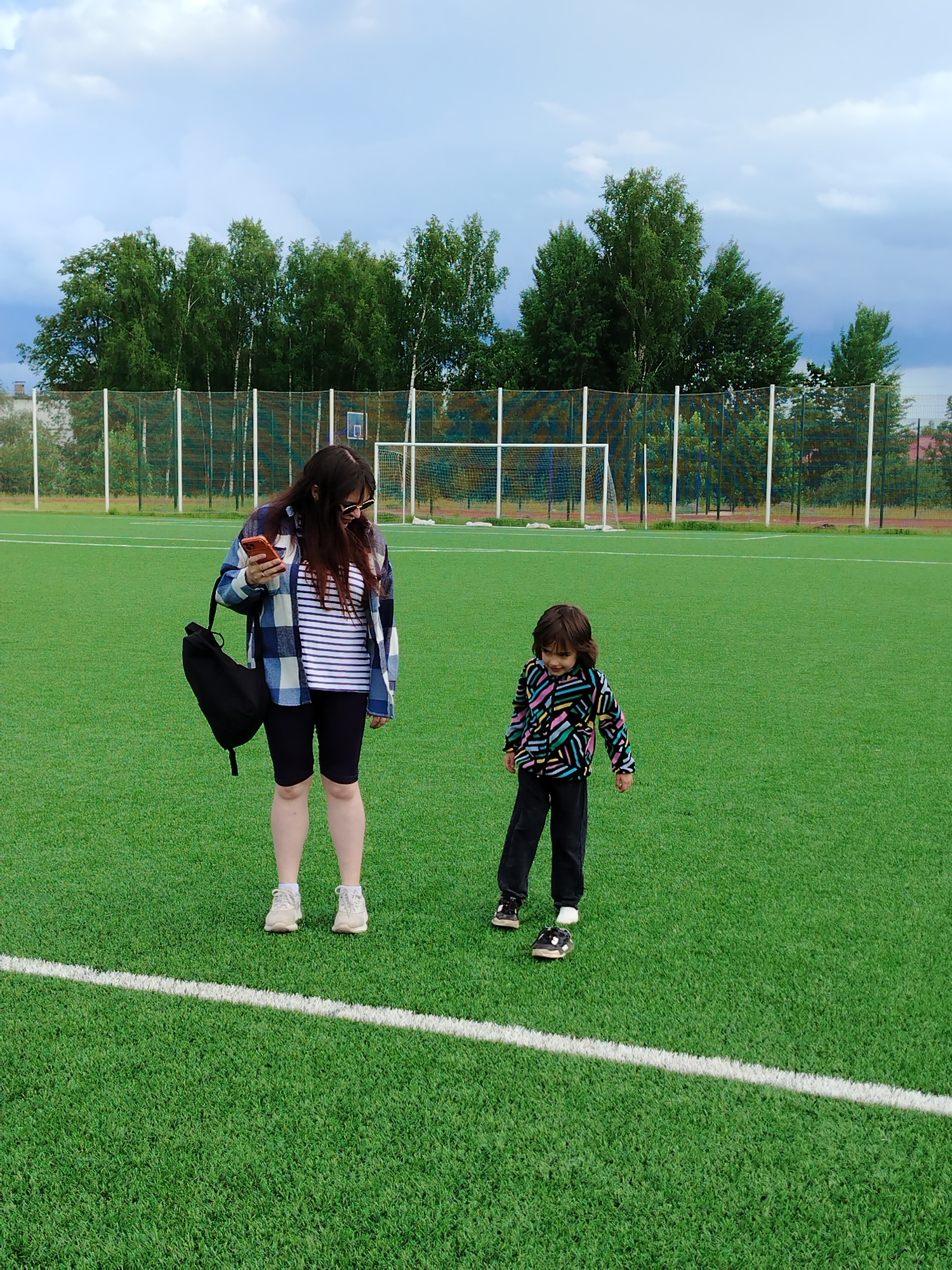 The image captures a woman and a child on a green artificial turf field in Noviy, Russia. The woman, wearing a blue plaid shirt, striped top, black shorts, and white sneakers, holds a smartphone in her hand, looking down at it. A black bag hangs from her shoulder. Beside her, the child is dressed in a colorful patterned jacket and dark pants, also wearing shoes.
In the background is a white soccer goal and a fence, partially obscured by green trees. Above, a cloudy sky is visible. The scene is shot in daylight. The white line on the soccer field lies between the adults legs and child's feet.