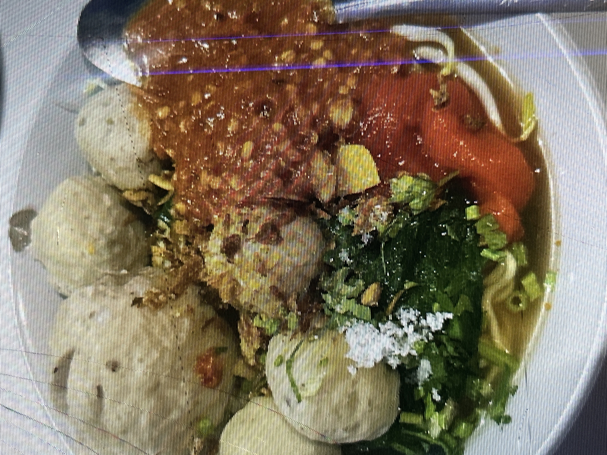 The image shows a close-up, overhead view of a bowl of food, likely a noodle soup dish with meatballs, served in a white bowl. The dish contains several large, light-colored meatballs, some smaller, textured meatballs, a generous portion of reddish-brown sauce, green leafy vegetables, chopped green onions, and what appears to be shredded white coconut or cheese. Thin yellow noodles are visible at the bottom of the bowl. A portion of a silver spoon is visible on the right side of the frame, dipping into the sauce. The lighting is overhead and appears somewhat uneven, with some glare. The image quality is degraded by horizontal lines, suggesting it may have been taken with a low-resolution camera or on a screen. There is no visible indication of location in Depok, Indonesia, or any people, actions, or interactions.