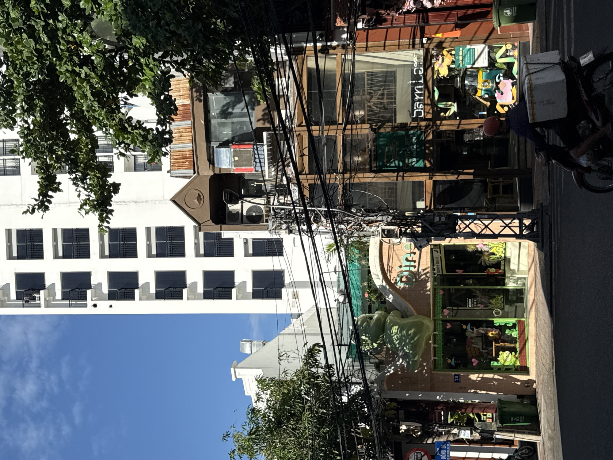 The image shows a street scene in Nha Trang, Vietnam. A tall white building with multiple windows and balconies dominates the background. In front of it, there are several shops and businesses, some with vibrant storefronts decorated with cartoon characters and colorful signs. One sign reads "Dino's Gelato." Another sign appears to be for a Thai restaurant. A motorcycle rider with a red helmet is visible in the foreground, carrying a white box on the back of their vehicle. The street is paved, and there are power lines and a traffic light visible. Trees with lush green foliage are present, casting shadows on the scene. The sky is blue with some clouds. The overall impression is that of a busy urban environment with a mix of modern architecture and local businesses.