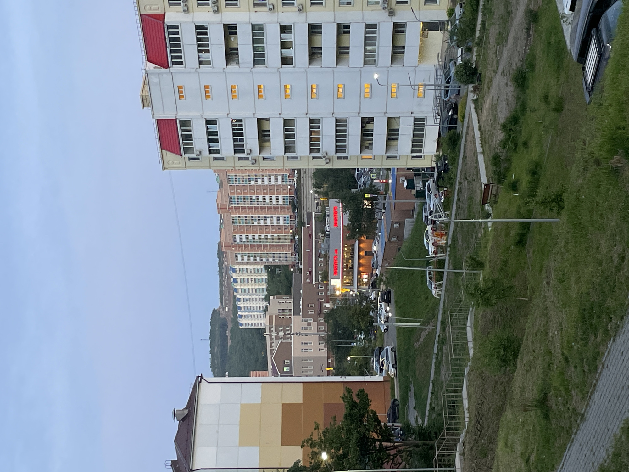The image shows an urban scene in Vladivostok, Russia. Multiple buildings of varying heights and architectural styles are visible, including a tall, white apartment building with a red roof. A street runs between the buildings with parked cars and streetlights. In the distance, a series of buildings descends towards a slightly elevated green space. The sky is overcast. The store sign "HOSTIP'S" is visible. A set of metal stairs is also visible.