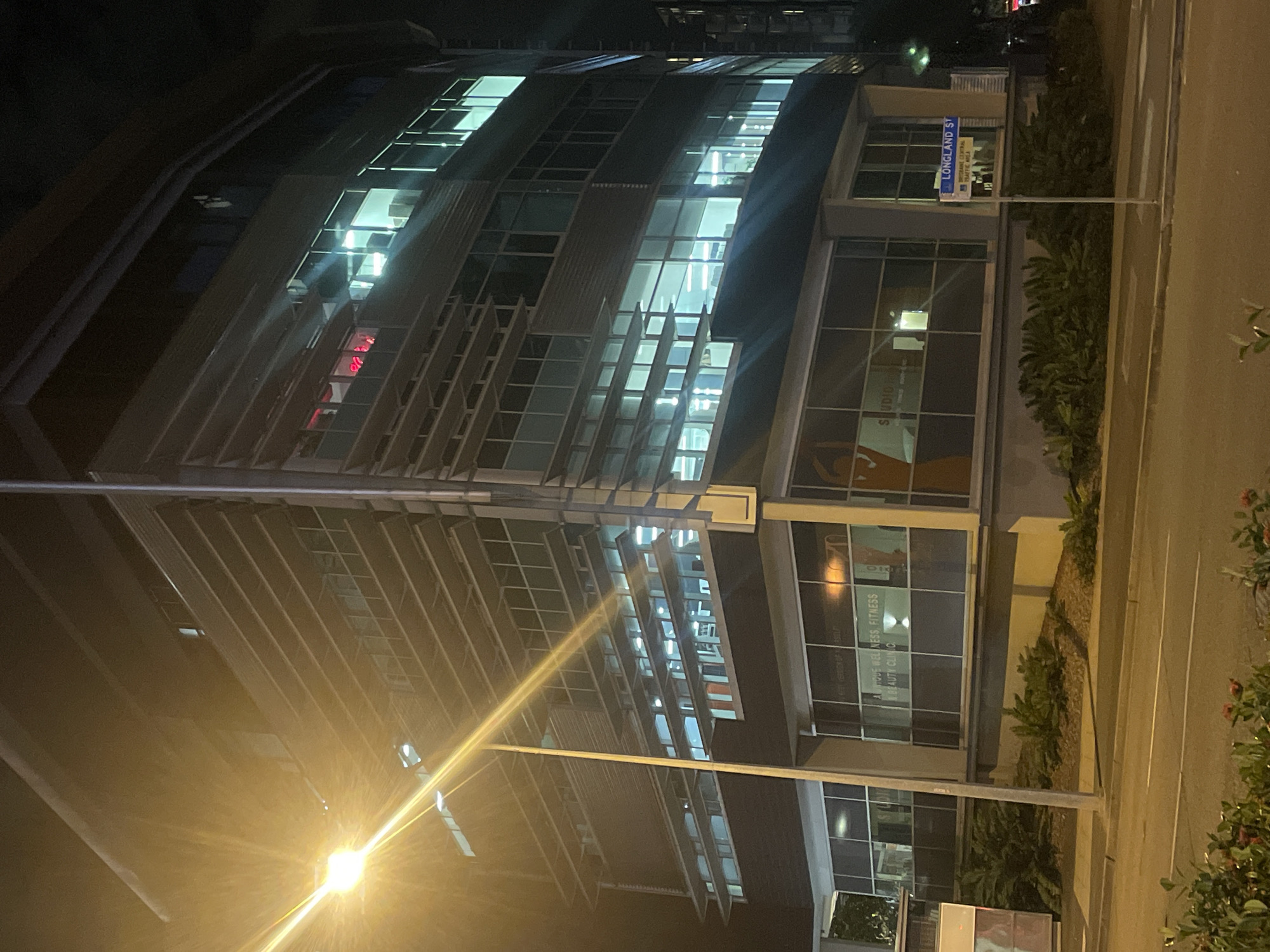 The image shows a multi-story building at night, illuminated by streetlights and interior lights. The building is made of glass and metal, with horizontal louvers on the exterior. On the ground floor, there are large windows with signage indicating a "STUDIO" and "ATTUNE WELLNESS, FITNESS & BEAUTY CLINIC." A street sign reading "LONG LAND ST" is visible on the right side of the image. The street in front of the building is paved and empty, with some vegetation in the foreground. The overall scene suggests an urban environment in Brisbane, Australia, likely a commercial or office building. There are no people visible in the image.