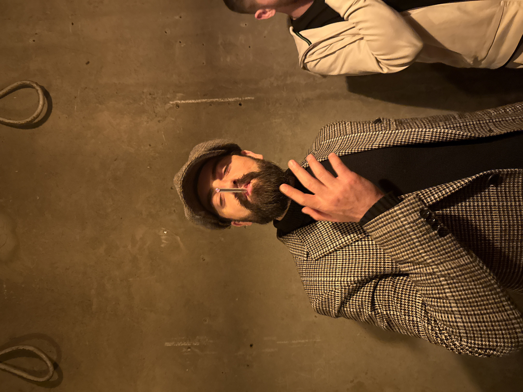 The image shows two men indoors with a dim, warm lighting. The man in the foreground wears a newsboy cap, a black turtleneck sweater, and a checkered brown and black blazer. He has a beard and is holding a cigarette in his mouth. His left hand is raised. Another man is visible in the upper right corner, wearing a light-colored jacket with black accents. The floor is a plain concrete surface, and a rope-like object is visible in the upper left and lower left corners. The location is Yerevan, Armenia.