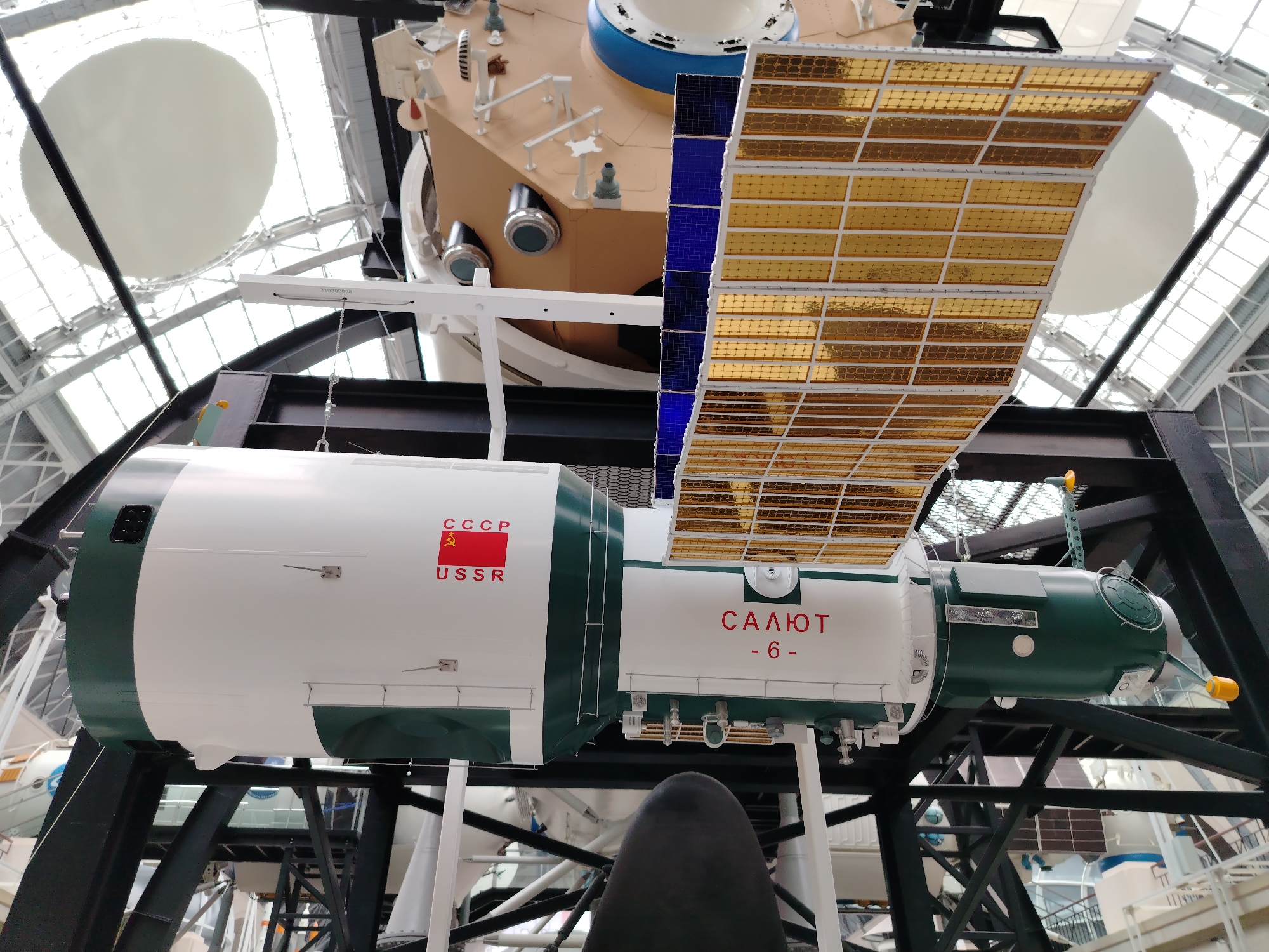 This media file captures a model of the Salyut 6 space station suspended within a large indoor exhibition space. The space station model is predominantly white and dark green, with a large solar panel array attached, featuring numerous golden cells. Text visible on the side of the station reads "СССР USSR" with a red flag and hammer and sickle emblem, and "САЛЮТ - 6 -". The station is mounted on a black metal support structure. In the background, the ceiling of the building is visible, featuring a large, circular white object, possibly part of another exhibit or the building's architecture, and a glass-paneled roof allowing natural light to enter. The overall setting suggests a museum or science exhibition, likely in Moscow, Russia, given the presence of the Salyut space station, a Soviet-era program. No people are visible in the image, and the activity is static, focusing on the display of the space station model. The lighting appears to be a mix of natural light from the roof and artificial interior lighting, suggesting it is daytime.