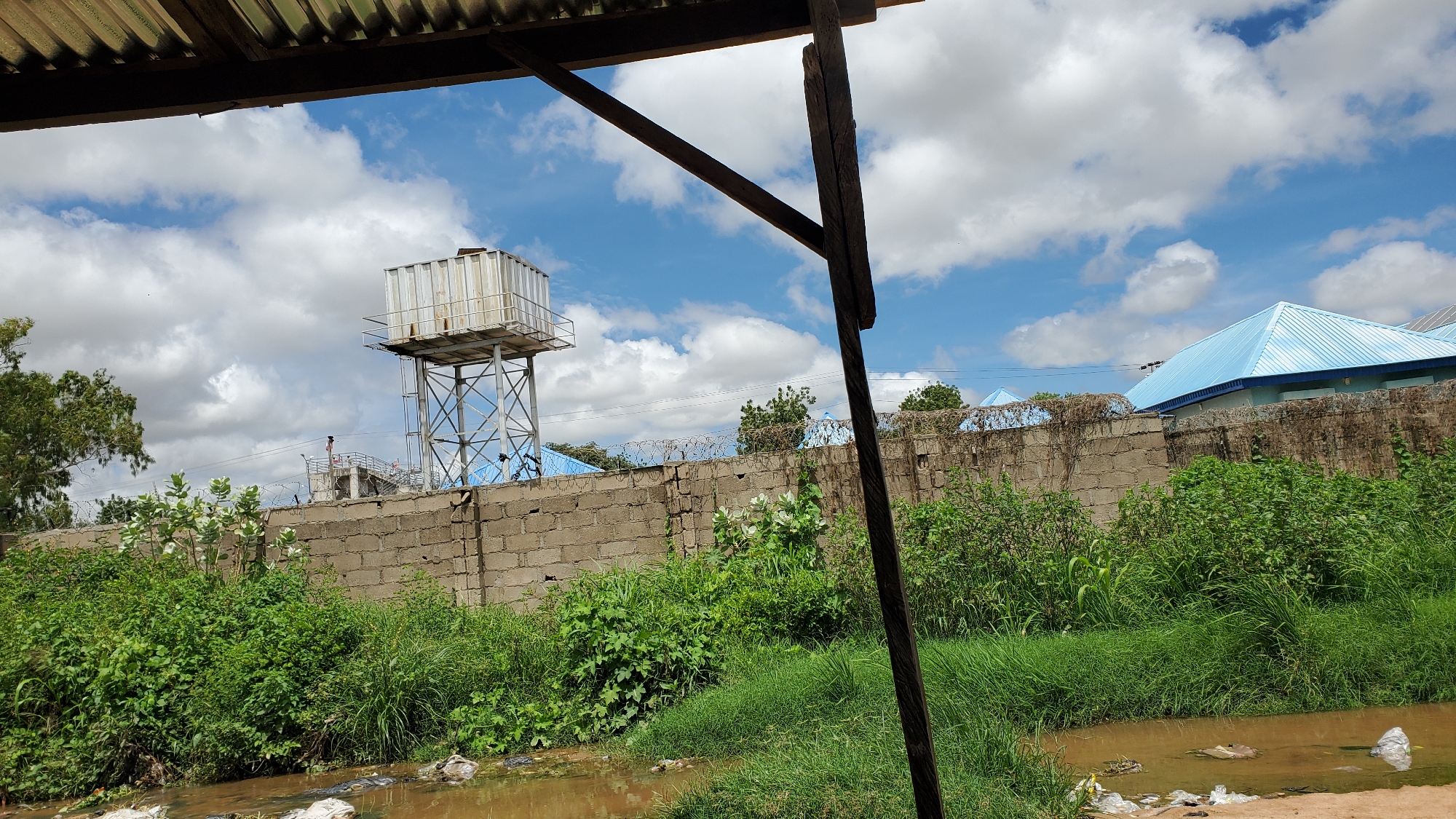 The image captures a view of a residential or industrial area in Jalingo, Nigeria, under a partly cloudy sky. In the foreground, a body of muddy water with visible debris, including plastic bags, suggests poor sanitation. A concrete wall, topped with barbed wire, runs horizontally across the mid-ground, separating the foreground from what lies beyond. Behind the wall, lush green vegetation thrives. Dominating the left side of the mid-ground is a tall metal water tower with a rectangular tank at the top. To the right of the water tower and beyond the wall, the brightly colored blue roofs of several buildings are visible, indicating ongoing construction or recently completed structures. A wooden beam protrudes from the upper left corner, framing the scene, and another wooden post is prominent in the right foreground, adding to the rustic and potentially unfinished feel of the environment. The overall scene depicts a blend of essential infrastructure, such as the water tower, with signs of neglect and urban development characteristic of some areas in Jalingo.