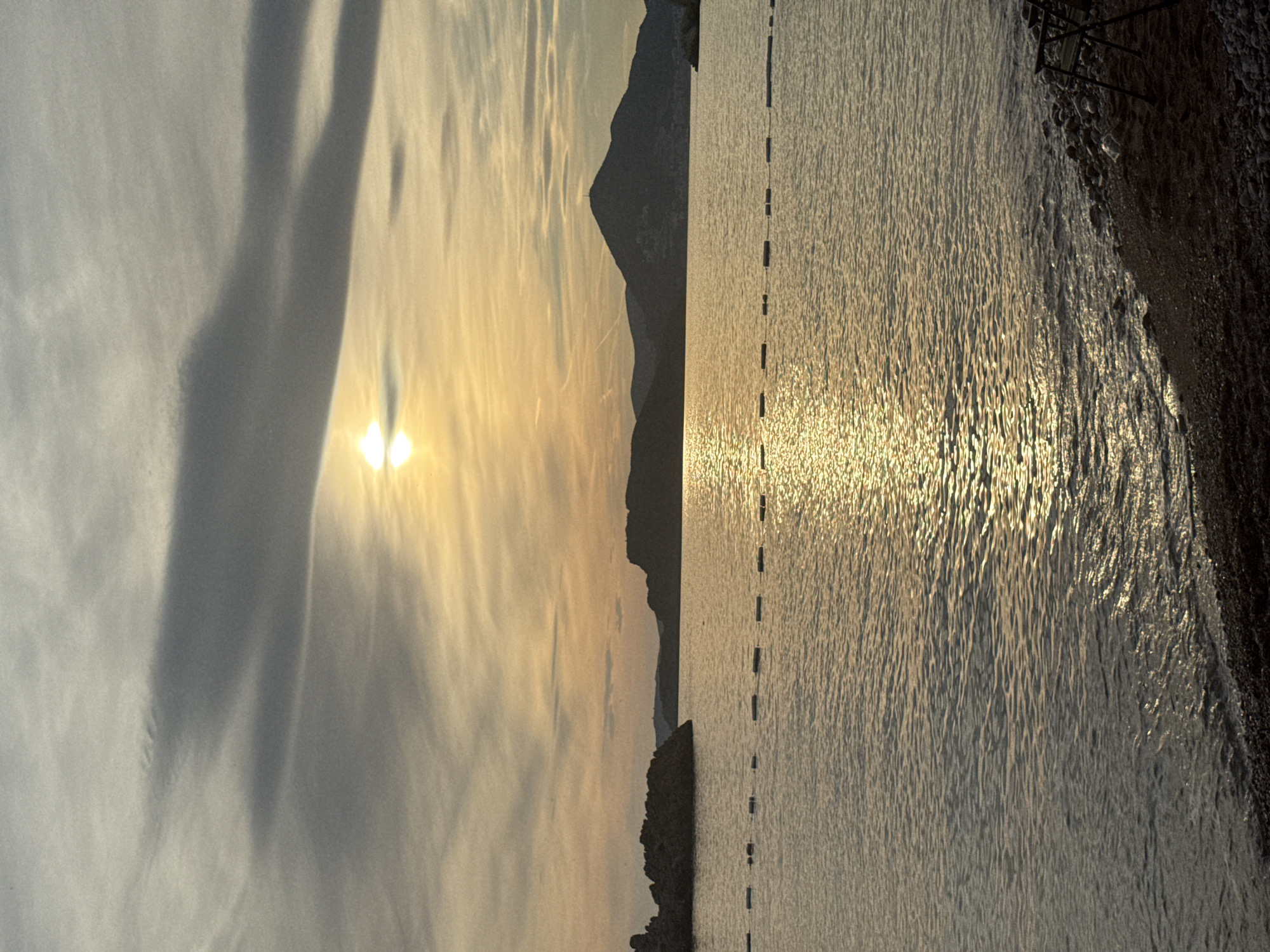The image depicts a serene sunset scene over the Adriatic Sea, likely in or near Sveti Stefan, Montenegro. The sun is partially obscured by wispy, horizontal clouds, casting a warm, golden glow across the water and sky. The sea is calm, with gentle ripples reflecting the sunlight. In the distance, silhouetted hills and mountains form the horizon, with a prominent peak featuring a small structure, possibly a tower, at its summit. On the left side of the frame, a rocky outcrop juts into the water. A line of white buoys floats on the surface, marking a boundary in the sea. The foreground shows a pebble beach with a dark, sandy area. Two chairs are visible on the right side of the beach, suggesting a place for relaxation and enjoyment of the view. The overall atmosphere is peaceful and tranquil, characteristic of a coastal sunset.