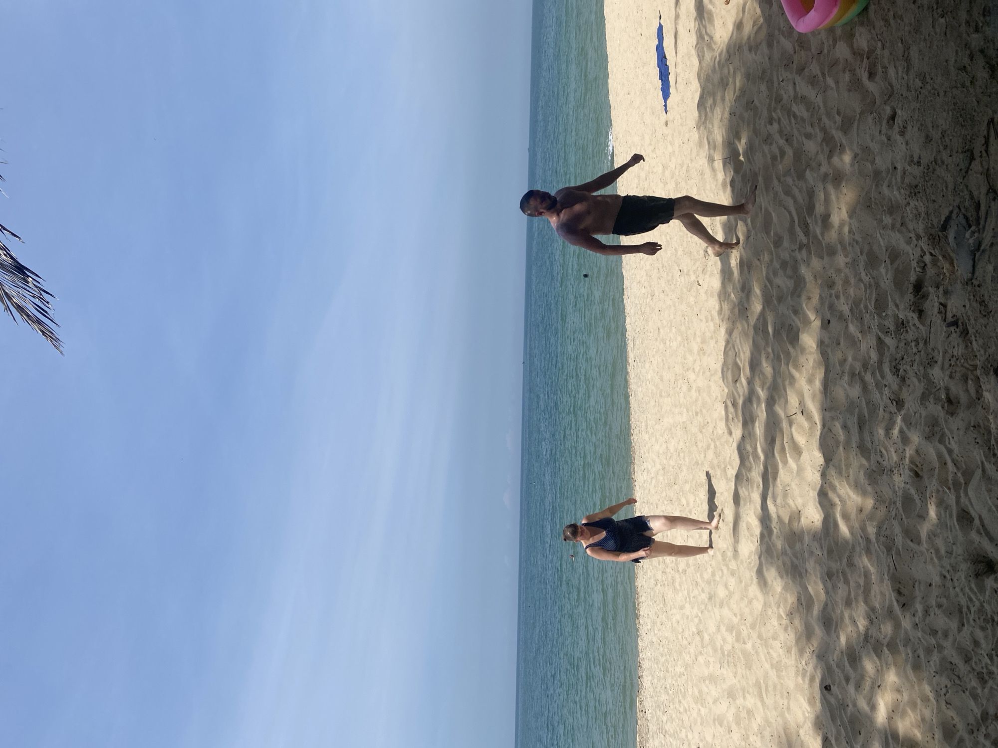This image depicts a beach scene in Phu Thuy Ward, Vietnam, with two individuals on the sand. On the left, a woman wearing a dark blue patterned swimsuit walks towards the foreground, her head down. To her right and slightly further back, a shirtless man in black swim shorts walks in the same direction, looking towards the woman. The ocean stretches out behind them, a calm expanse of blue-green water under a clear, pale blue sky. The sandy beach is lightly shaded by the presence of palm fronds from the upper left corner of the frame. In the foreground, shadows cast by unseen objects create dappled patterns on the sand. A blue towel lies on the sand to the right of the man, and part of a colorful inflatable toy is visible in the lower right corner. The scene is captured on a bright, sunny day.