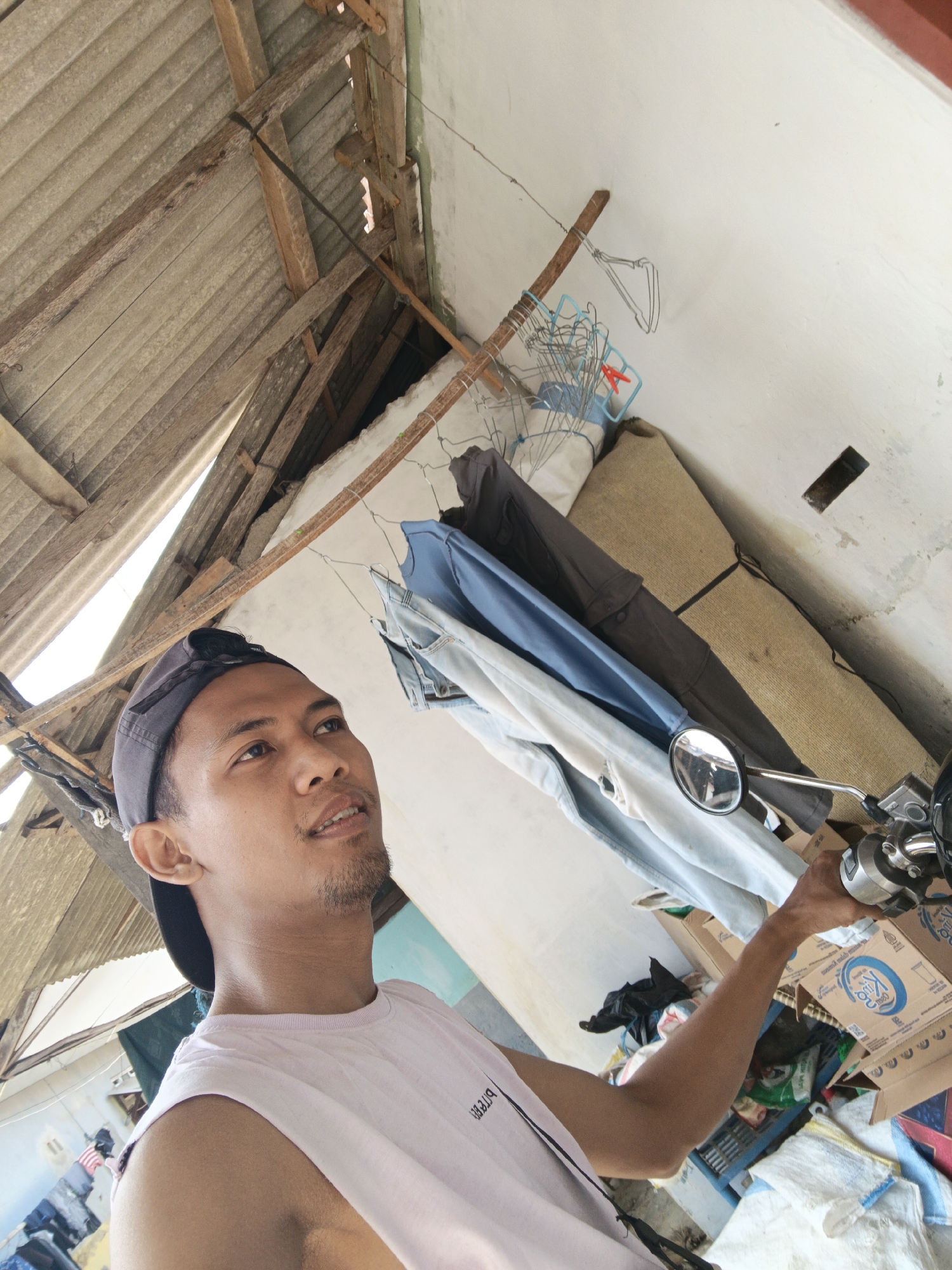A low-angle, medium shot captures a young man in Singolatren, Indonesia, posing for a selfie. He is wearing a light pink sleeveless shirt and a dark baseball cap backward. He is standing next to a motorcycle, with his right hand on the handlebars. The background reveals a cluttered, unfinished space. Overhead, a corrugated metal roof is supported by weathered wooden beams. Clothes hang on a wire strung across the wall, and various items, including bags, boxes, and what appears to be a rolled-up mat, are piled on the floor. The overall impression is of an informal, possibly rural, setting undergoing construction or in a state of disarray.