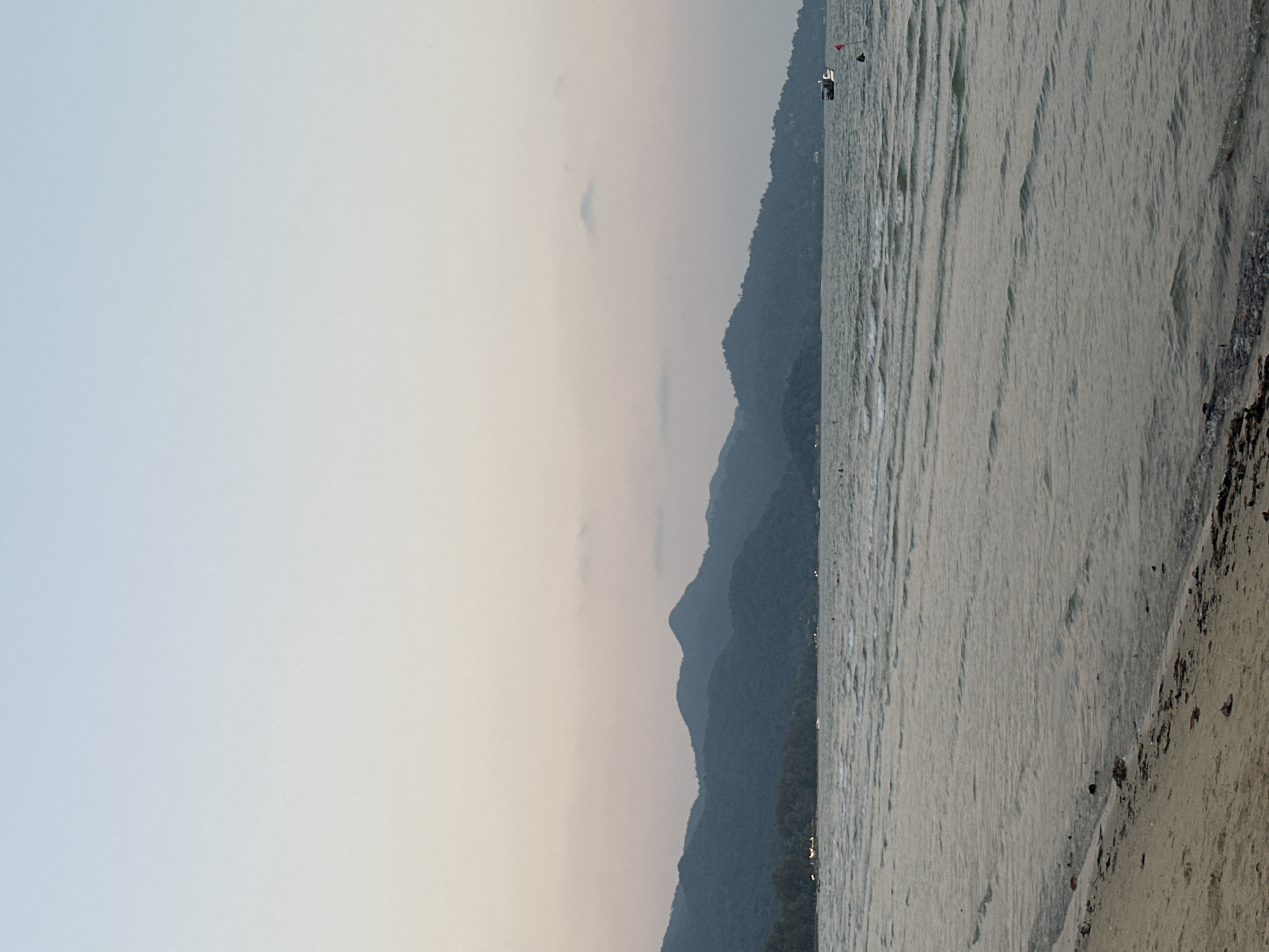 A scenic view in Baan Klong Prao, Thailand, shows the coastline meeting a silhouetted mountain range under a hazy sky. The sea is gently rippled, extending from the shore toward the distant mountains. A sandy beach borders the edge of the image. There is a boat located further out in the water.