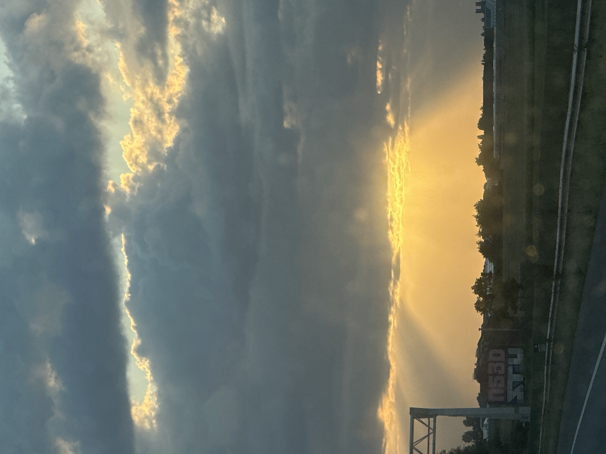The image captures a dramatic sunset over Rijpwetering, Netherlands. Heavy, dark gray clouds dominate the upper half of the frame, with shafts of golden sunlight breaking through in several places. A large band of bright, luminous clouds stretches across the horizon, casting a warm glow over the landscape. Below this, a field of dark green grass extends towards a row of trees and some buildings. To the left, a concrete overpass structure is visible, and on a building behind it, red and white graffiti reading "N53D STU" is present. The foreground shows a portion of a paved road and a grassy verge. The scene suggests a late afternoon or early evening setting.