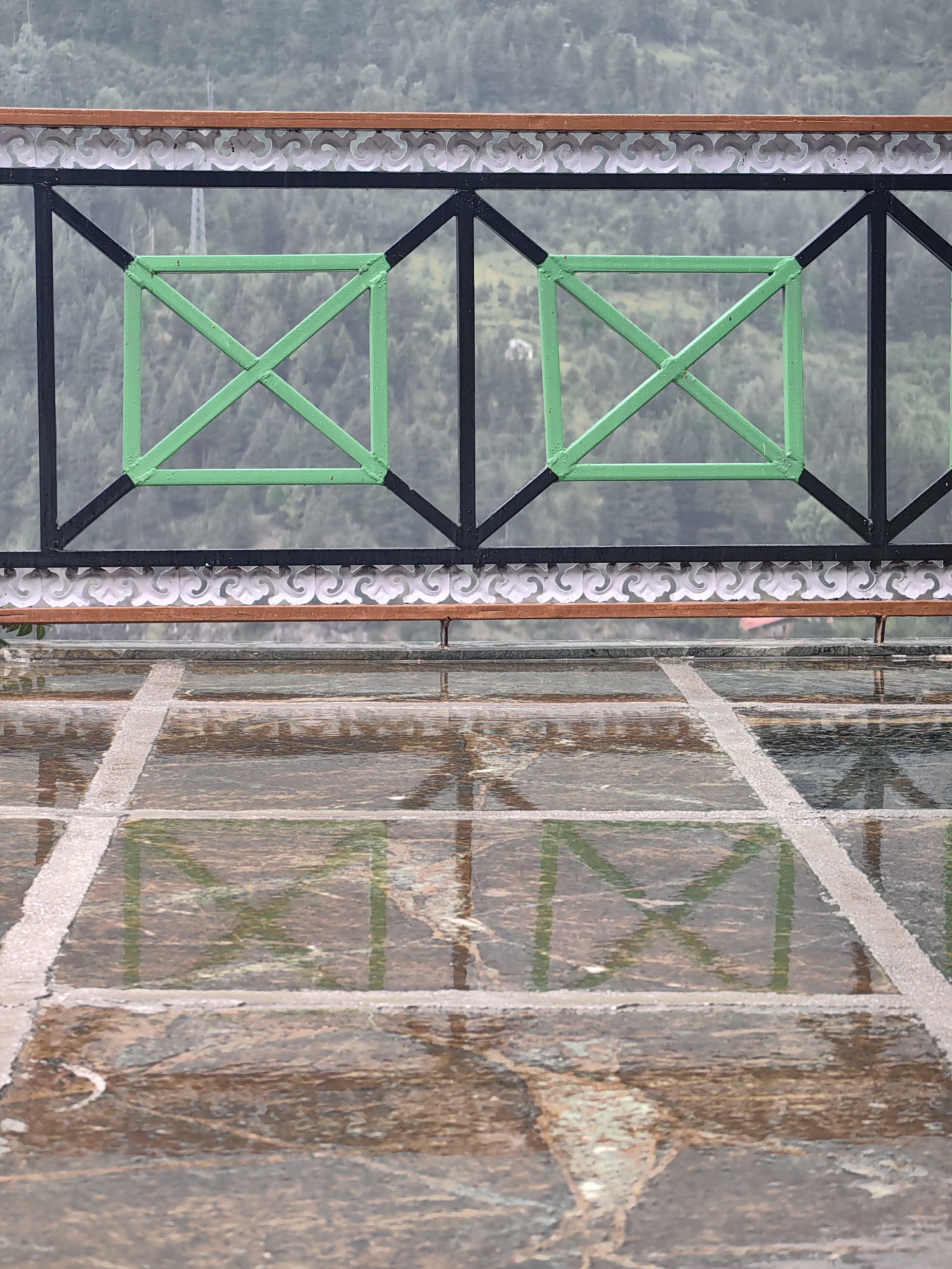 The image depicts a balcony or viewing platform in Vashisht, India, overlooking a misty, forested mountainside. A black metal railing with decorative white trim and a wooden top rail separates the platform from the view. Within the railing, two prominent green square frames with crisscrossing supports are visible. The platform floor is made of large, dark stone tiles, wet from rain, creating reflections of the railing and the landscape. The reflections on the wet floor are a significant feature, showing a distorted, watery mirror image of the scene above. The overall atmosphere is subdued due to the mist and rain, with muted colors dominating the scene. There are no people or animals visible in the image.