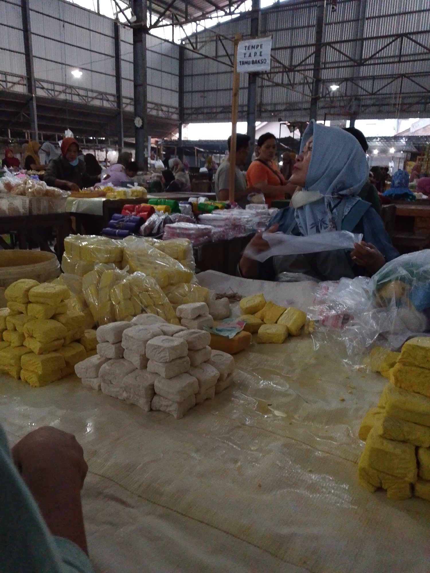 The image depicts a busy indoor market scene in Pepedan, Indonesia. In the foreground, stacks of yellow, rectangular tofu are prominently displayed on a tarp-covered surface. To the left and right of the tofu, there are further stacks of similar yellow tofu, some wrapped in plastic. In the middle ground, to the right of the yellow tofu, there are stacks of white, cubed tofu. Behind the tofu, a woman wearing a light blue headscarf is sitting and holding a piece of clear plastic. She is looking upwards and to the right. She is partially obscuring a display of various packaged goods, including brightly colored items. Other market-goers are visible in the background, engaged in transactions or browsing. The market appears to be covered by a large, corrugated metal roof supported by a metal structure. A sign with Indonesian text, "TEMPE TAPE TAHU BAKSO," is visible hanging from a wooden pole in the upper right section of the image. The overall impression is of a vibrant, local food market with a focus on tofu and other food items.