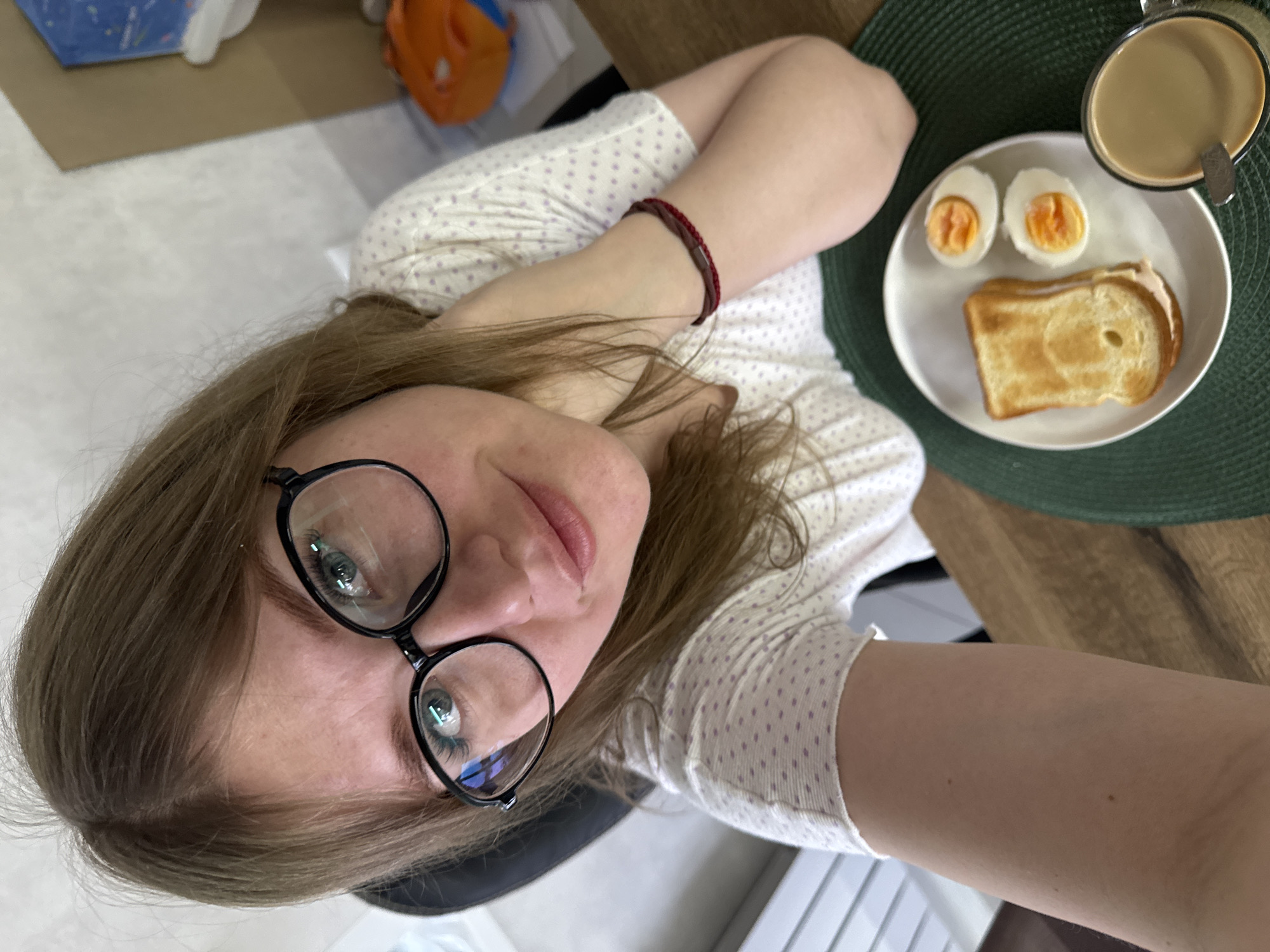 In an indoor setting within Gornyi (Dvurechenskoe S/P), Russia, a light-skinned individual with light brown hair and wearing black-framed glasses looks directly into the camera, capturing a high-angle self-portrait. The individual wears a white short-sleeved top with small purple polka dots and a dark red bracelet on the right wrist, with their right hand positioned near the neck/chin.
Before the individual, on a wooden table surface, a meal is arranged on a dark green woven placemat. A white ceramic plate holds two slices of toasted bread, appearing as a sandwich, alongside two hard-boiled eggs cut in half, revealing bright orange yolks. Adjacent to the plate, a clear glass mug contains a light brown beverage, possibly coffee or tea with milk, with a metal spoon inserted.
The background consists of a light-colored wall with a subtle textured pattern. To the left, part of a dark chair back is visible. To the right, a cardboard box and a partial view of an orange object are discernible. The scene is illuminated by ambient indoor light.