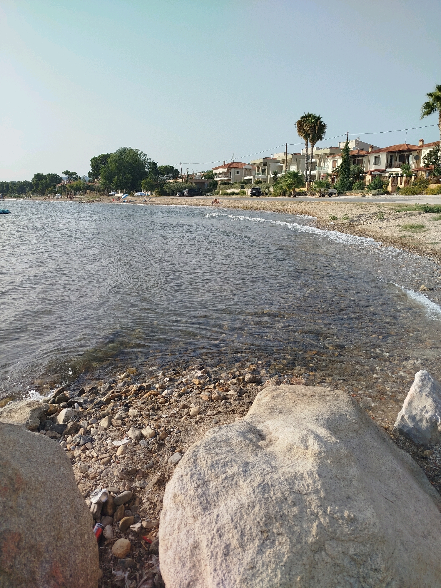 The image depicts a coastal scene in Sithonia Municipal Unit, Greece. The foreground features large rocks and a pebbled shoreline with some scattered debris, including a crushed aluminum can. The shallow, clear water gently laps at the shore, revealing the rocky seabed. In the mid-ground, a sandy beach is visible, lined with trees and residential buildings. Several palm trees stand out among the vegetation. A road runs parallel to the beach, with a few parked cars. Further in the background, more trees and buildings are visible under a clear, pale sky. There are a few people scattered along the beach, some sitting or lying down, and a couple of small umbrellas are set up. The overall impression is a quiet, sunny day at a seaside settlement.