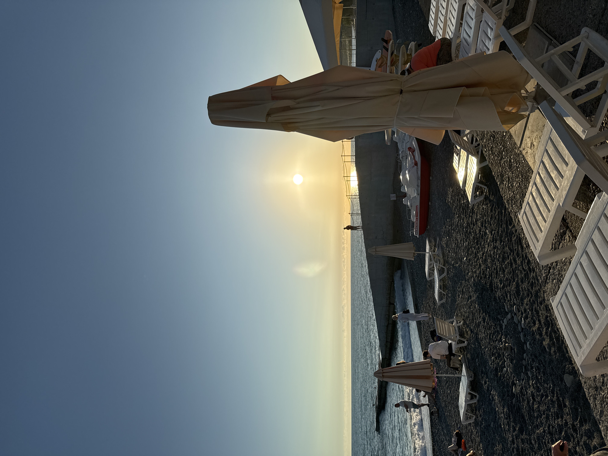The image depicts a beach scene in Sochi, Russia, during sunset. The sun is low on the horizon, casting a warm glow over the water and the pebble beach. In the foreground, several empty white lounge chairs are arranged, some with folded umbrellas. Further back, people are visible near the water's edge, with waves gently lapping the shore. A concrete pier extends into the sea on the left, and a few pedal boats are parked on the sand. A single figure stands on the pier, silhouetted against the setting sun. The sky is a clear, pale blue, transitioning to a soft yellow and orange near the horizon. The overall atmosphere is serene and peaceful.