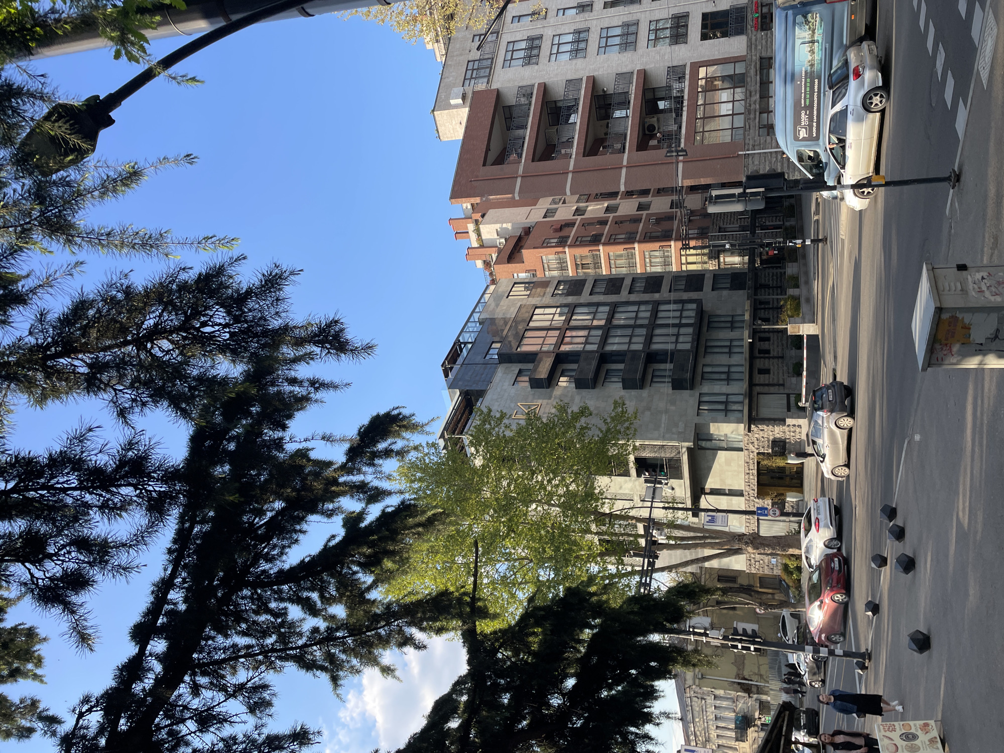 The image captures a vibrant urban street scene in Tbilisi, Georgia, under a clear, bright blue sky with scattered clouds. The perspective is slightly upward, with a large, dark coniferous tree canopy dominating the left foreground and a dense deciduous tree near the center.
**Subjects:**
The scene is framed by multi-story city buildings of varying architectural styles, featuring numerous windows, balconies, and some with modern facades. The street runs horizontally across the middle and right side of the image, with several cars parked or waiting. These include a white Subaru, a light blue Ford Transit-style van, a maroon hatchback, a white hatchback, and a dark grey car. Street furniture such as traffic lights, streetlights, and dark hexagonal bollards line the street. Two women are visible on the sidewalk in the bottom right, one wearing a dark jacket and jeans, the other in a black top. Another person can be seen inside the white Subaru.
**Activity or Event:**
The image depicts everyday city life. Cars are present on the road, either parked or in motion, suggesting traffic flow. People are walking on the sidewalk, and a street stall indicates commercial activity. The overall impression is one of a bustling city environment.
**Notable Details:**
The time of day appears to be mid-morning or afternoon, indicated by the strong sunlight and shadows cast by the buildings and trees. The weather is sunny and clear. A key location cue is the light blue van, which has the text "MAGRO CITY" and "GEORGIA" along with a phone number starting with "+995 32", clearly indicating its Georgian origin and confirming the Tbilisi location. Some buildings have air conditioning units visible on their exteriors. One building features a distinctive stylized 'Z' or similar emblem on its facade.
**Visible Text:**
The most prominent legible text is on the side of the light blue van: "MAGRO CITY", "GEORGIA", and a phone number that appears to be "+995 32 210 10 30". There are also graphics of food items (e.g., bread, vegetables) on a street stall sign, but no readable text.