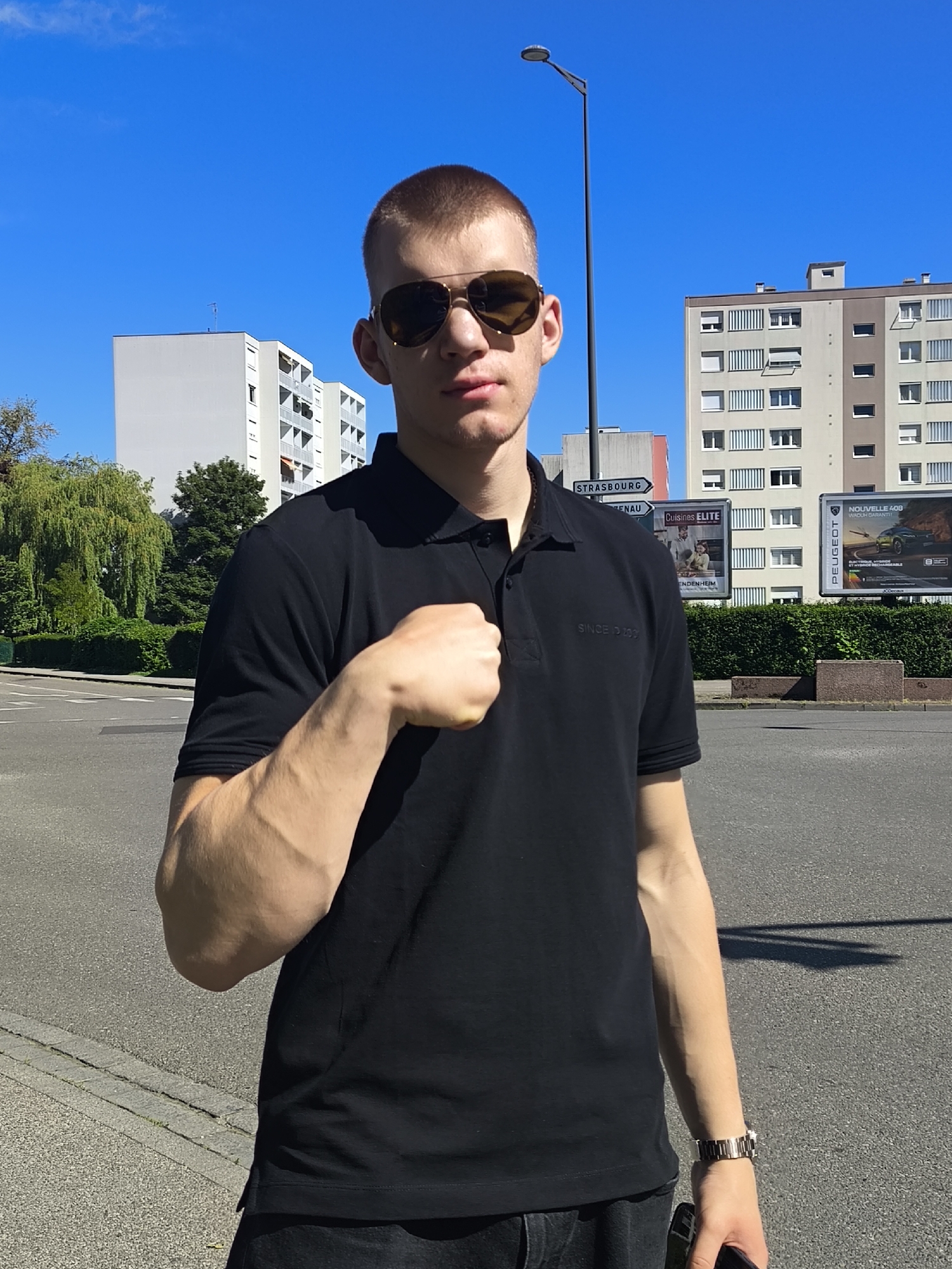 A young man stands centered in an outdoor urban setting in Hoenheim, France, under a clear blue sky. He has short, light brown hair and wears dark sunglasses, a black polo shirt with faintly visible text "SINCE 1993", and dark trousers. A silver-toned watch is on his left wrist, and he holds a smartphone in his right hand, which is partially obscured. His left arm is bent, with his fist lightly clenched and resting against his upper body, suggesting a confident or casual pose.
The background features several multi-story residential apartment buildings, typical of a European city. To the left, there is lush greenery including trees and hedges. The man stands on an asphalt road next to a sidewalk.
Notable details include the bright, sunny weather with strong shadows, indicative of midday or early afternoon. A tall street lamp is visible behind him. Location cues are prominent: a white road sign with a black arrow points left, displaying "STRASBOURG". Further back, two billboards are visible. The left billboard advertises "Cuisines ELITE" located "À MUNDOLSHEIM". The right billboard features a "PEUGEOT" advertisement for the "NOUVELLE 408", highlighting its "ELECTRIQUE, HYBRIDE" (Electric, Hybrid) features and "VOITURE GARANTIE" (Guaranteed Car). The overall scene depicts a typical street environment in a residential area.