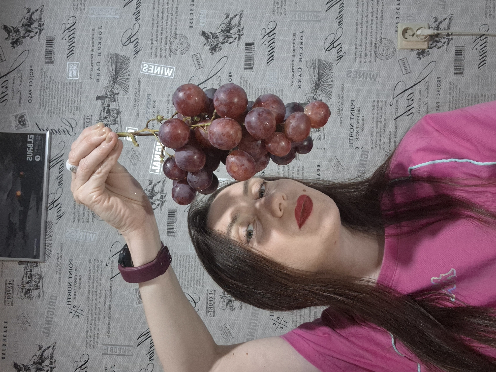 A woman in a pink shirt holds a bunch of red grapes above her head. She is wearing a purple watch on her left wrist and a ring on her left ring finger. She has dark hair, light skin, and red lipstick. The background is a grey wallpaper with wine-themed text and images. A black object is visible on the left side of the image. A power outlet is visible on the top right of the image. The image is rotated counter-clockwise.