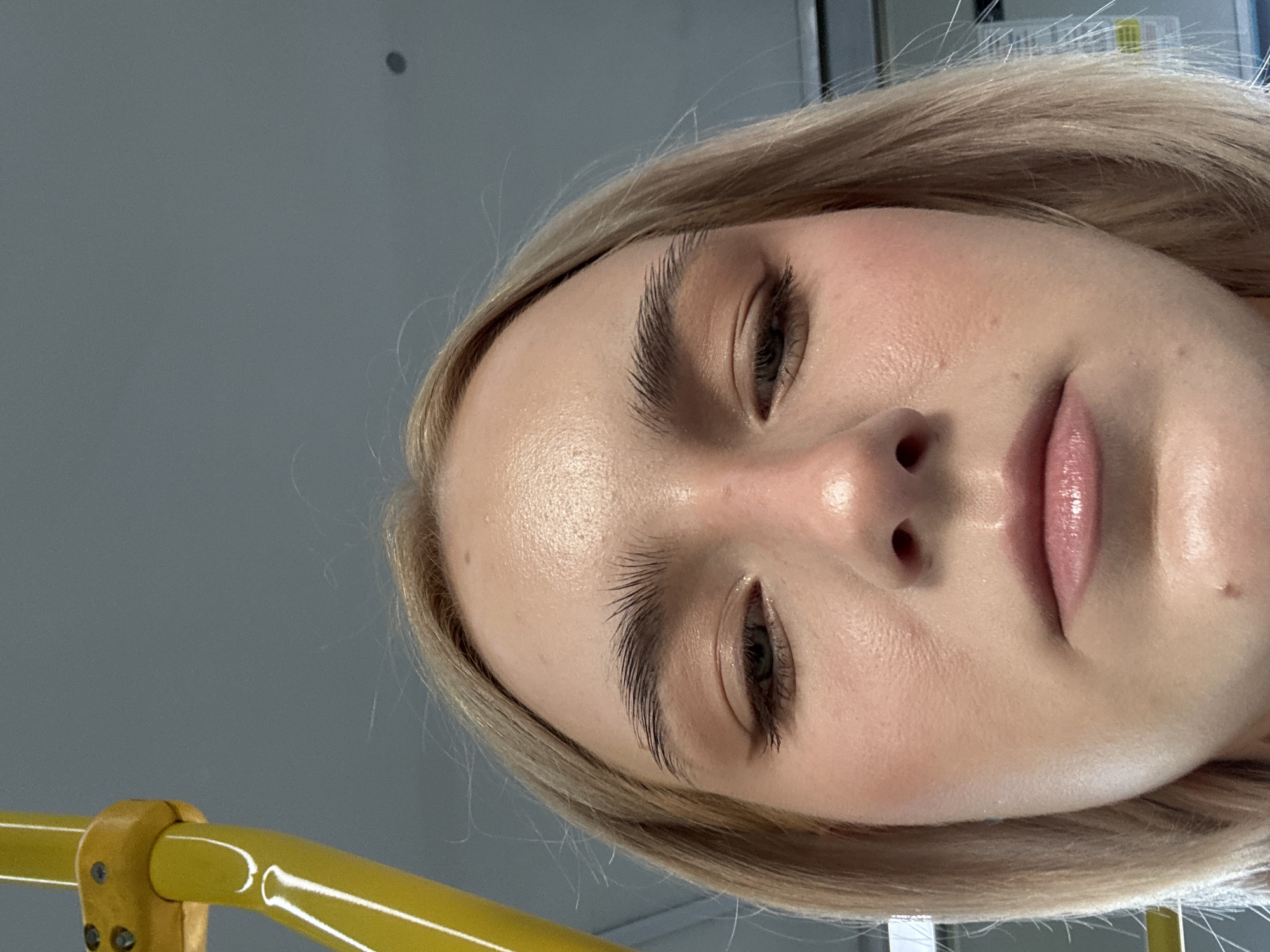 A close-up, portrait-style image captures the face of a person, likely female, with light blonde hair. The individual's head is tilted to the right, and the face is angled slightly upwards, suggesting a selfie perspective. She has fair skin with a subtle sheen on her forehead and cheeks, and her eyebrows are dark and well-groomed. Her eyes are partially visible, looking slightly upwards, and her lips are a soft pink, possibly enhanced with gloss. There's a hint of light blush on her cheeks.
In the foreground, across the bottom left of the frame, is a prominent bright yellow, glossy cylindrical bar or handrail, possibly metal or plastic, with visible attachment points. The background consists of a plain, out-of-focus gray surface, which could be a wall or ceiling, with a small, indistinct dark spot visible in the top left. The presence of the yellow bar suggests an indoor public setting, possibly a form of public transport like a bus or train. No text is visible in the image.