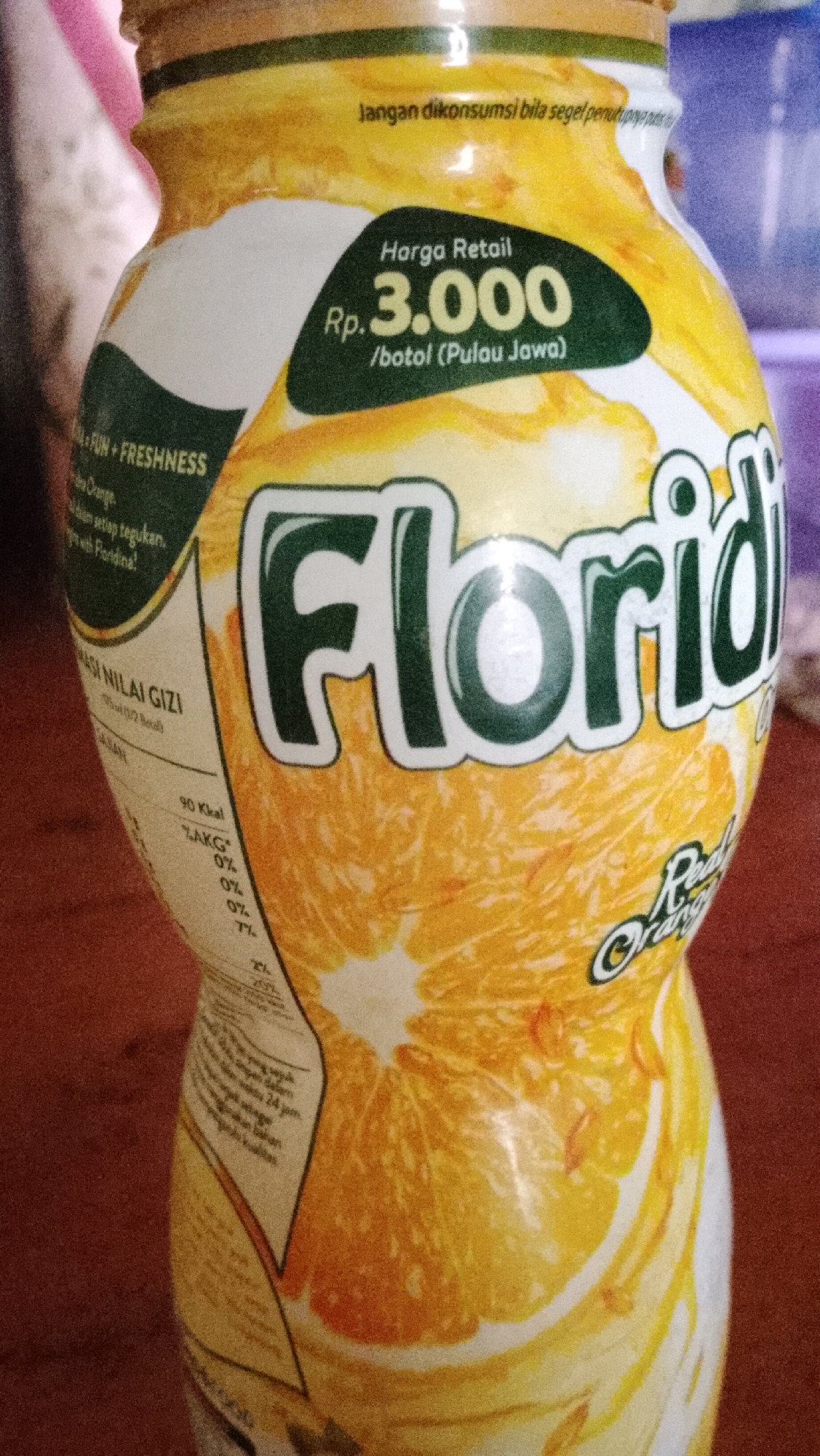 A close-up, eye-level, indoor shot shows a plastic bottle of Floridina brand orange juice. The bottle is predominantly white with a yellow cap and features a large, stylized illustration of an orange slice. The text on the bottle is in Indonesian. A green oval shape contains the price information: "Harga Retail Rp. 3.000 /botol (Pulau Jawa)", which translates to "Retail Price Rp. 3,000 /bottle (Java Island)". Other text includes nutritional information, slogans like "FUN + FRESHNESS", and a warning "Jangan dikonsumsi bila segel tertutup". The lighting is bright, casting a slight glare on the bottle's surface. The background is blurred, suggesting a domestic or retail setting. The specific location in Bojong Barat, Indonesia, is not determinable from the image.