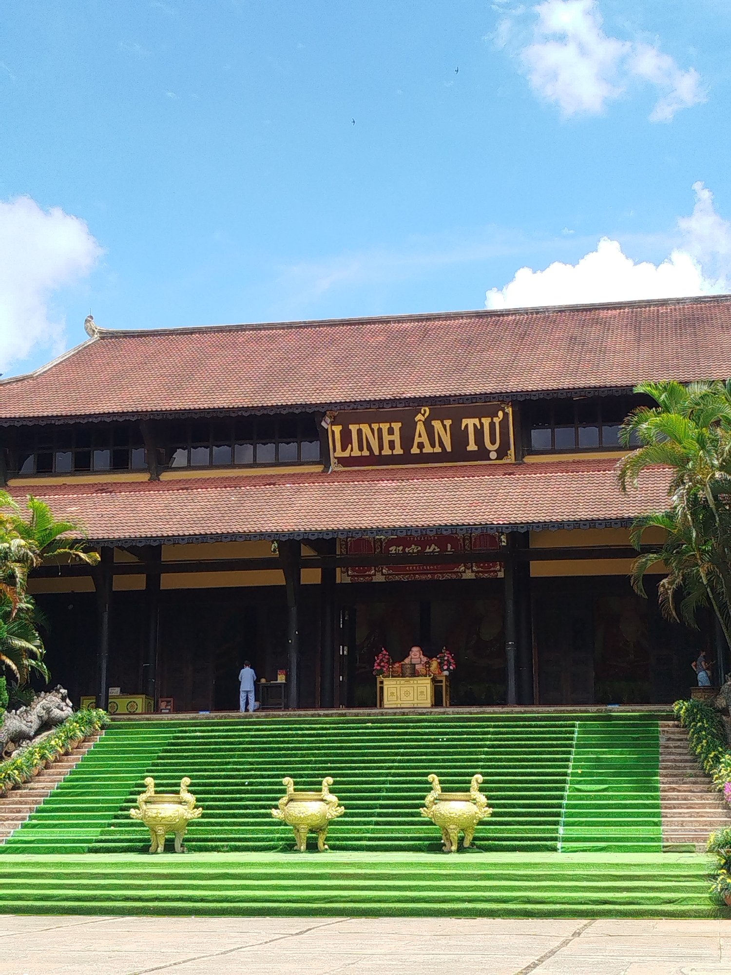 The image depicts the Linh An Tu Pagoda in Nam Ban, Vietnam. The pagoda is a multi-tiered structure with a brown tiled roof and dark wooden pillars. A large sign with the name "Linh An Tu" is displayed prominently on the facade. Wide green steps, adorned with three gold decorative pots, lead up to the entrance of the pagoda. A person in white clothing is standing near the entrance, while a statue of a smiling figure is placed on a gold stand within the pagoda. Lush greenery and palm trees surround the pagoda, with a blue sky and scattered clouds overhead. The overall scene presents a serene and architectural view of the Linh An Tu Pagoda.