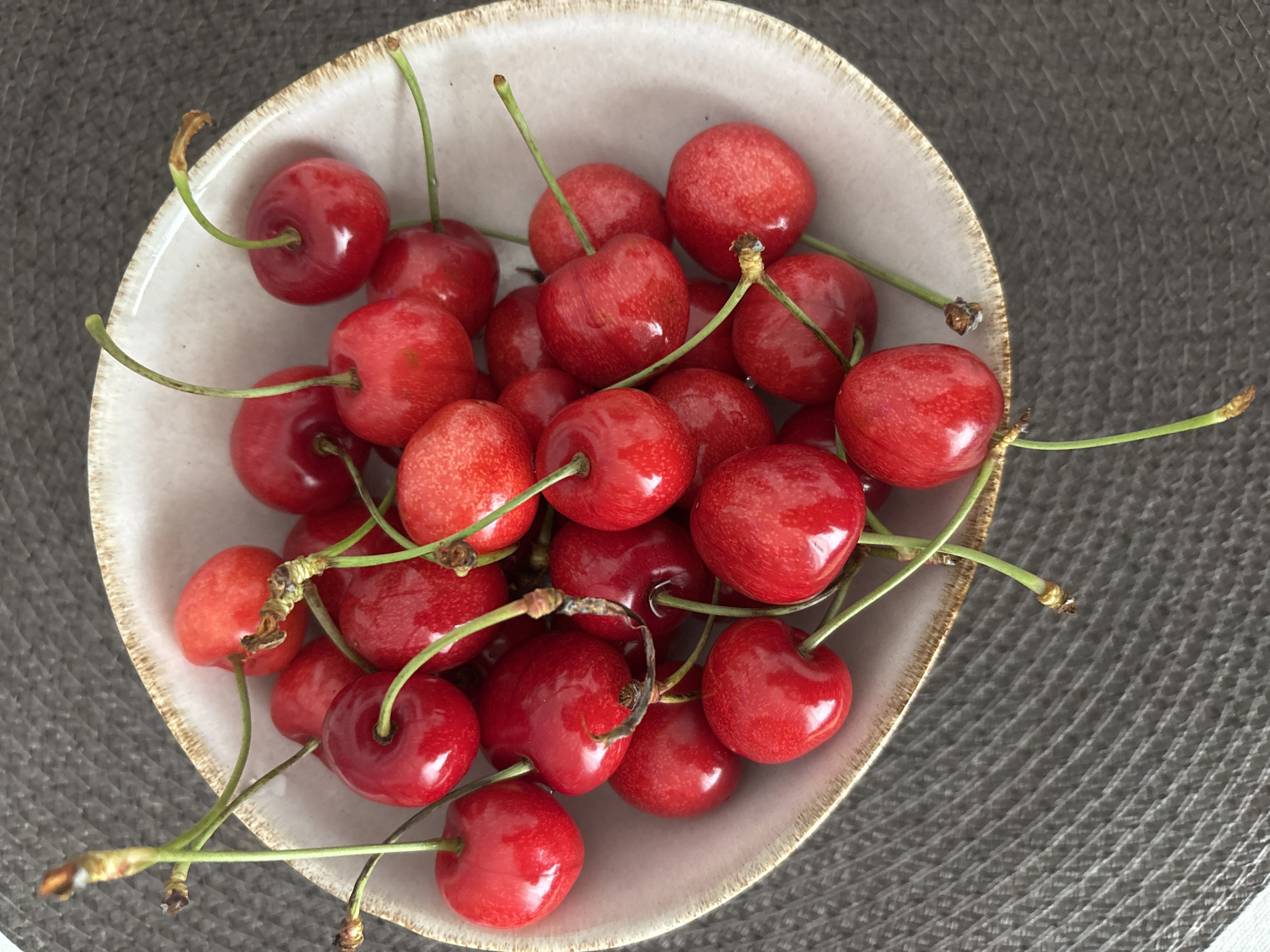 A bowl of cherries is pictured from directly above. The bowl is white with a beige rim and sits on a dark gray placemat with a woven texture. The cherries are bright red and many still have their green stems attached. The lighting suggests an indoor setting. No specific location in Saint Petersburg, Russia, can be identified from this image.