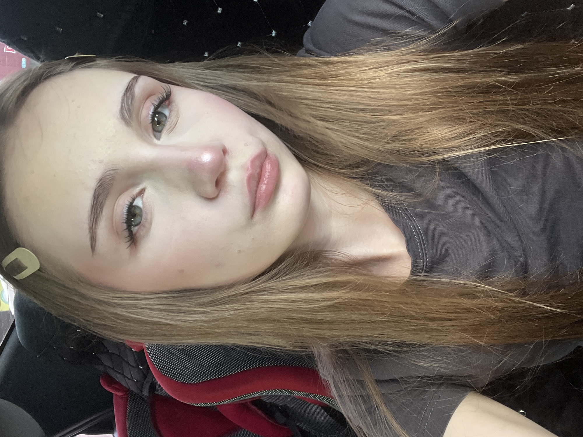 A young woman with blonde hair, wearing a dark gray t-shirt, is pictured from the chest up. She is sitting in what appears to be a car, with a red and black car seat visible behind her. She has light makeup on, including eyeshadow and lipstick. A square-shaped hair clip is visible in her hair. The background is dark, with some faint lights visible on the right side. The image was likely taken indoors, possibly in the city of Загородный, Russia, given the context.