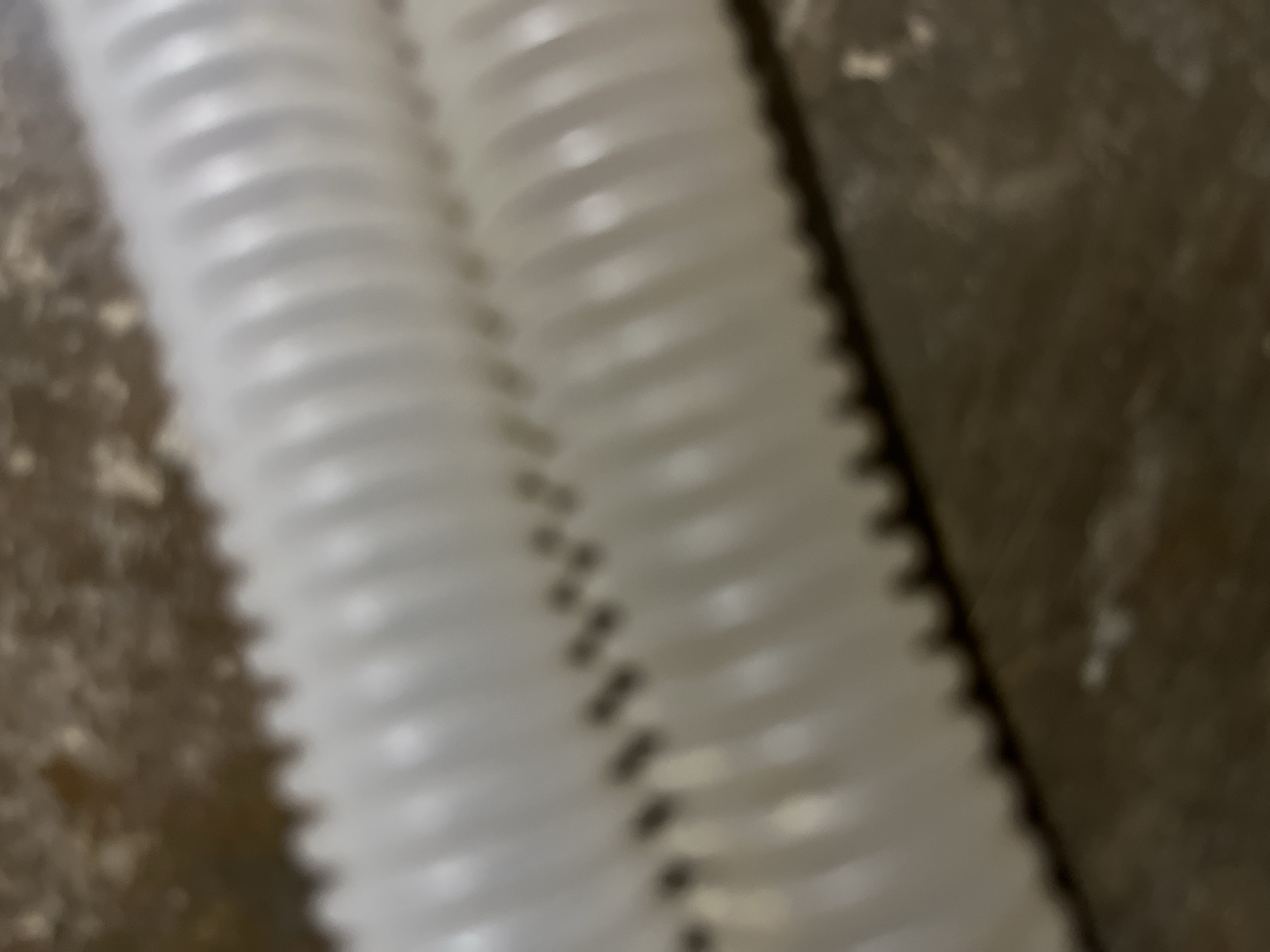 The image shows a close-up, blurred view of two white, corrugated plastic tubes lying horizontally on a textured, brown surface. The tubes are parallel and positioned diagonally from the bottom left to the top right of the frame. The surface they rest on appears to be a rough, possibly wooden or concrete material, with some lighter flecks scattered across it, suggesting dust or debris. The lighting is soft, casting subtle shadows and highlights on the ribbed texture of the tubes. The scene is indoors, and the environment suggests a workshop, storage area, or construction site. There are no people or discernible actions taking place. The image is devoid of any specific geographical markers that would indicate its location in Buleleng, Benuaraja, Indonesia.
