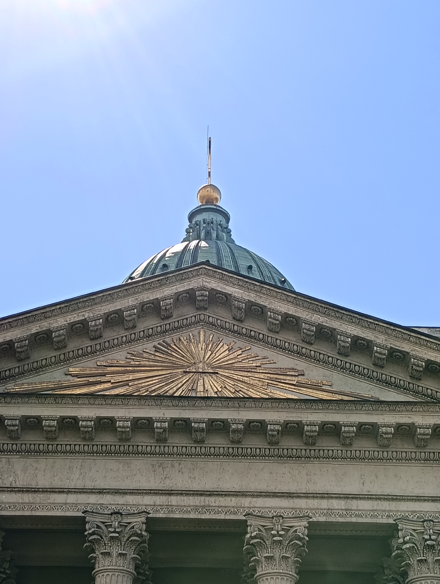 This is a low-angle, exterior photograph of the Kazan Cathedral in Saint Petersburg, Russia. The main subjects are the cathedral's dome and a section of its facade. The dome, a muted green with ornate architectural details, is capped by a golden sphere and a thin spire. Below the dome, the facade features a triangular pediment adorned with a golden, radiant sunburst motif. The building's stonework is intricate, with decorative moldings, corbels, and a frieze of circular ornamentation. Visible at the bottom of the frame are the tops of large, ornate columns, suggesting a colonnade. The sky is clear and bright blue, with a lens flare originating from the upper left corner, indicating it is daytime, likely mid-morning or afternoon, with sunny weather. The image conveys a sense of grandeur and architectural magnificence. No people are visible, and there is no discernible text in the image.