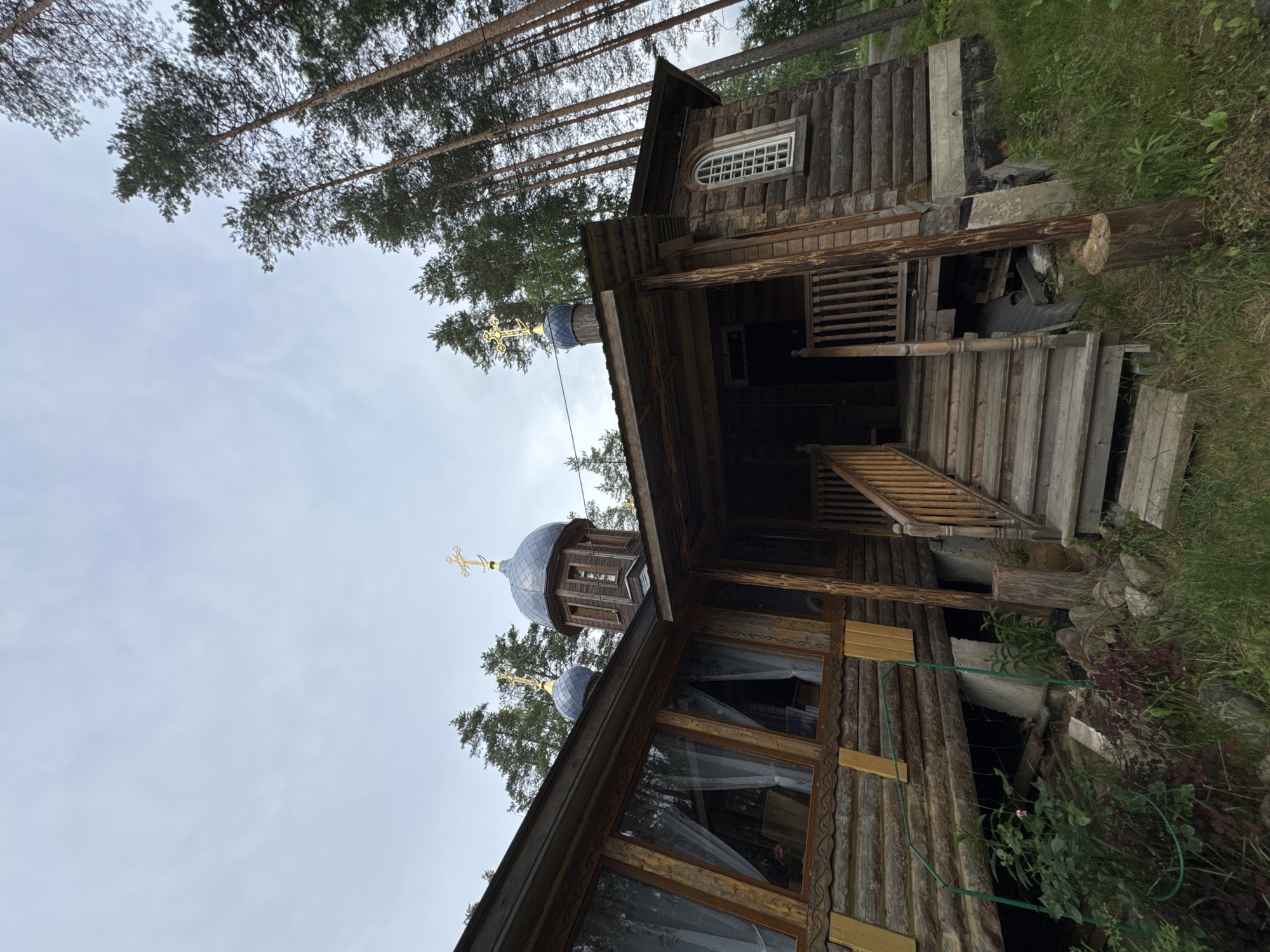 The image shows a wooden church in the village of Nenimäki, Russia. The church has two blue domes topped with golden crosses and a wooden facade. A wooden staircase leads to the entrance of the church. The church is surrounded by tall pine trees and lush green grass. The sky is overcast, and the overall atmosphere is peaceful and serene.