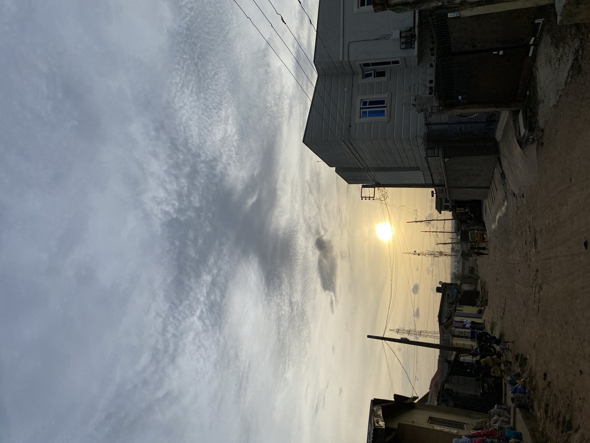 A ground-level shot, taken from a street, depicts a sunlit area in Lagos, Nigeria. The sky, predominantly on the left, features scattered clouds. Power lines crisscross the scene. On the right, buildings line the street. A three-story building with rectangular windows is in the foreground. Farther down the street are smaller buildings and shops with people gathered around. In the distance, towers stand near a road where a yellow vehicle and pedestrians are visible.