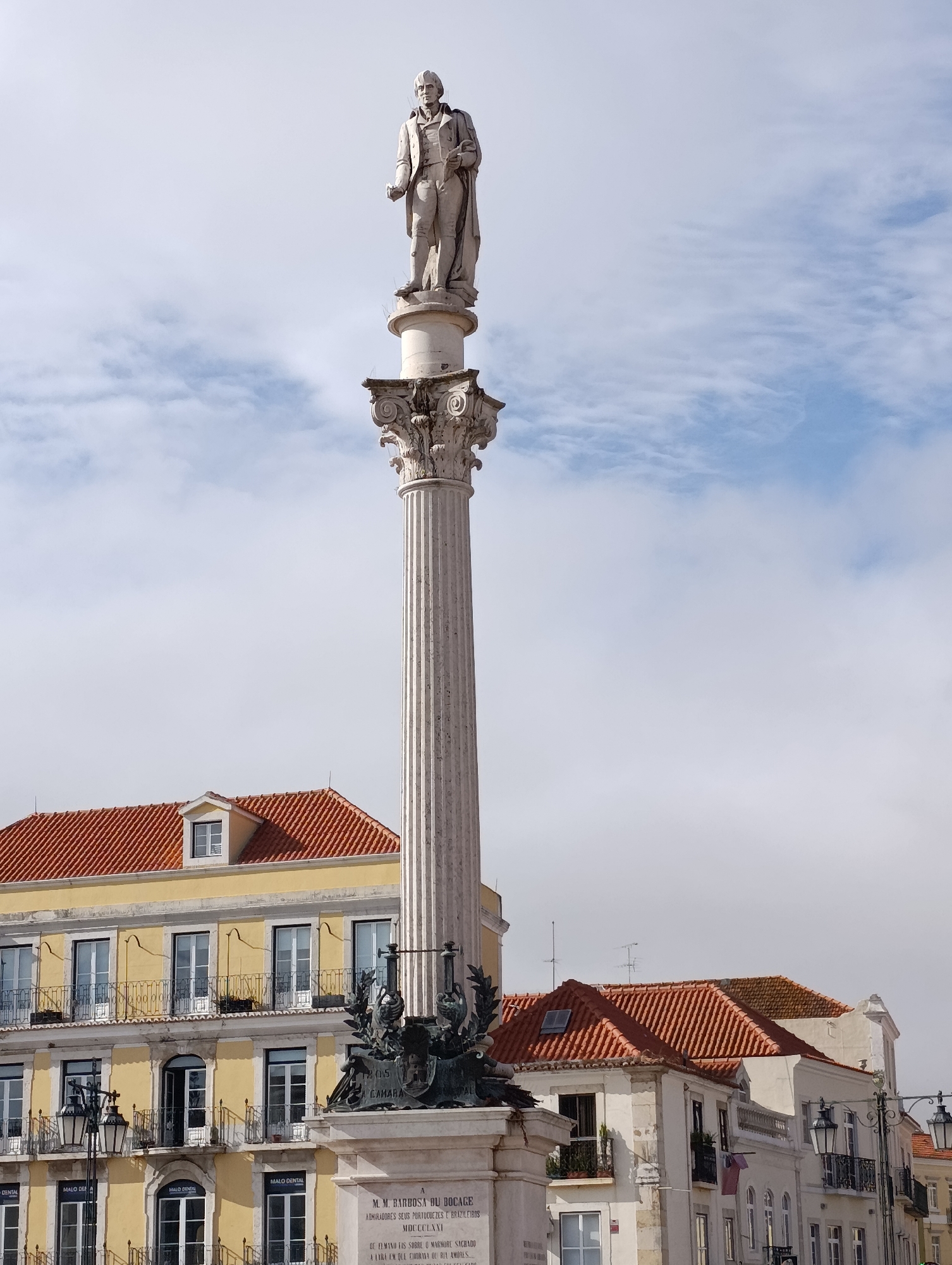 The image depicts the Monument to Bocage, a prominent landmark in Setúbal, Portugal. The monument consists of a tall, fluted column topped with a statue of the poet Manuel Maria Barbosa du Bocage. The statue portrays Bocage in formal attire, holding a book or scroll. The column itself is adorned with decorative elements near its capital.
At the base of the monument, a sculptural group featuring allegorical figures and inscriptions is visible. The inscription "1805 CAMARA" is partially visible on the monument, indicating a connection to the year 1805 and the city's council.
Behind the monument, buildings with characteristic red-tiled roofs are seen. One building, painted yellow, features multiple stories with balconies and windows. Another building, to the right, is off-white with similar architectural features. Streetlights with multiple lamps are present in the foreground. The sky above is partly cloudy with patches of blue. The scene captures a historical monument within an urban setting.